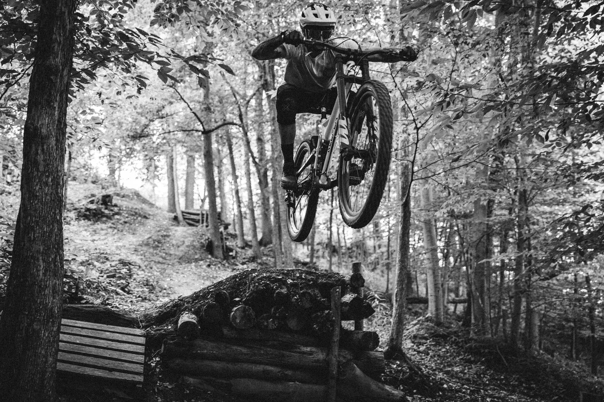 Mountain biker in mid-air over a log jump in a forest, black and white photograph.