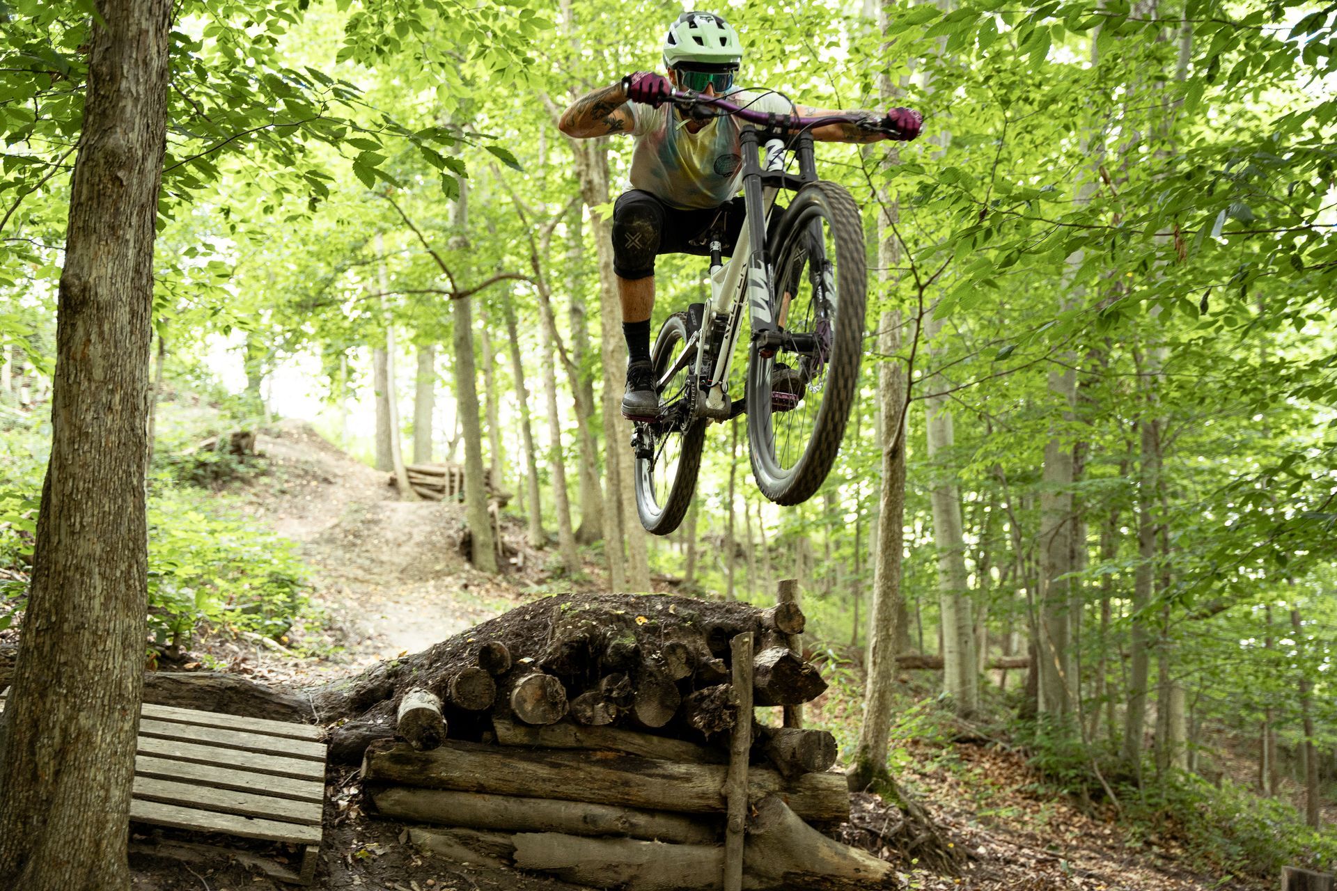 Mountain biker in mid-air over a wooden jump in a forest, wearing a helmet and protective gear.