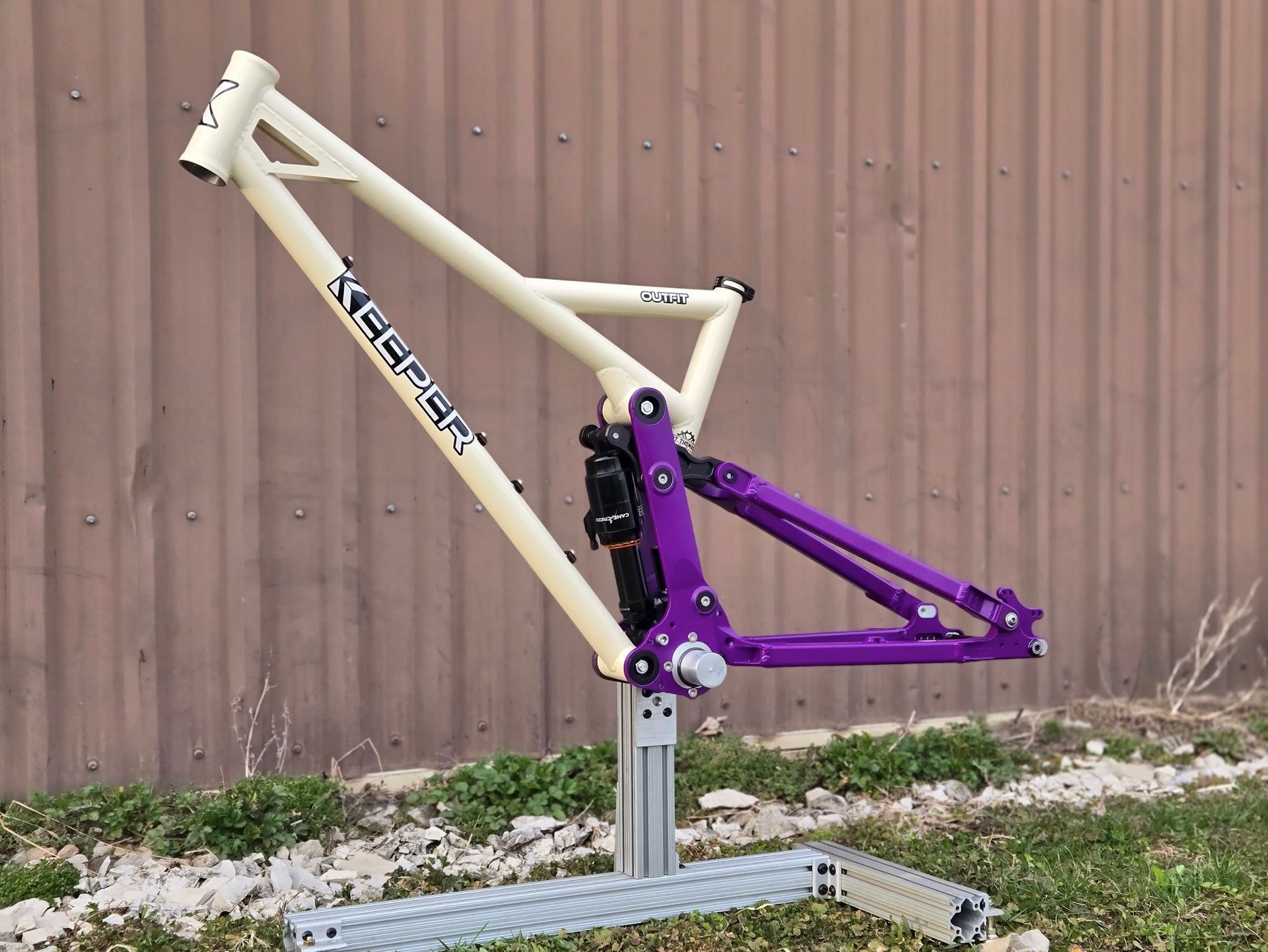 Cream and purple mountain bike frame on a silver stand, outdoors against a brown corrugated metal wall.