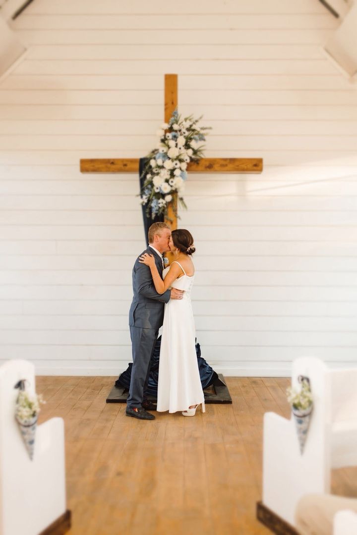 Newlyweds kissing in front of a wooden cross decorated with flowers, inside a white chapel.