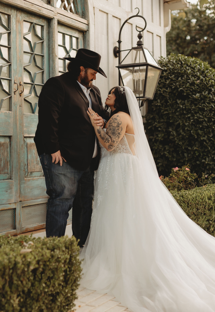 Couple in wedding attire: woman in gown looks up at man in cowboy hat; outdoor setting.