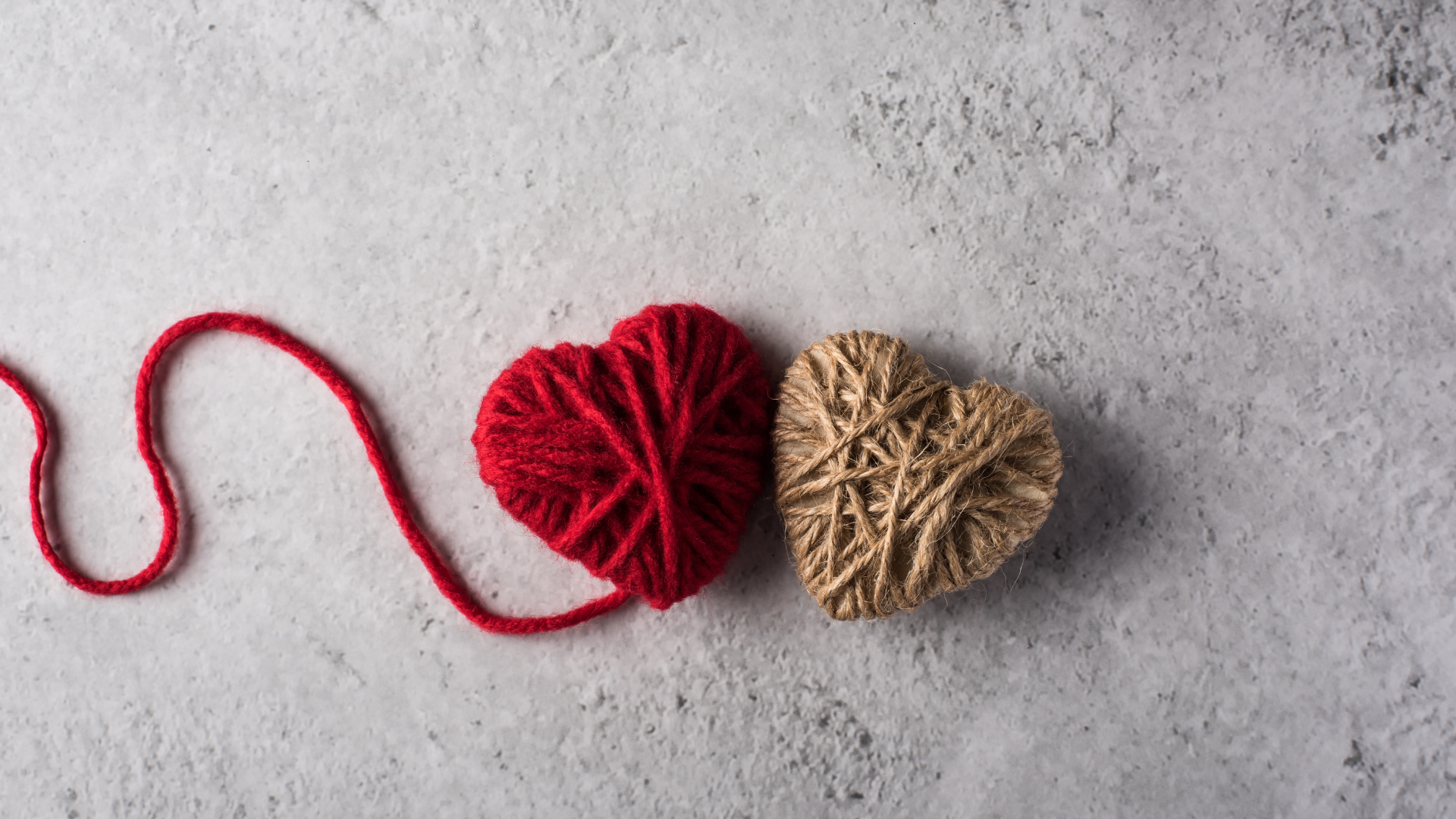 Deux cœurs en laine, l'un rouge et l'autre beige, reliés par un fil rouge, sur un fond gris texturé.