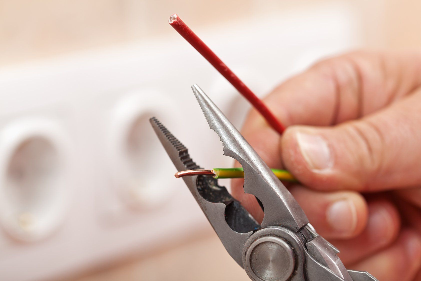 Pliers peeling copper wires - closeup on electrician hands, wall sockets in the background