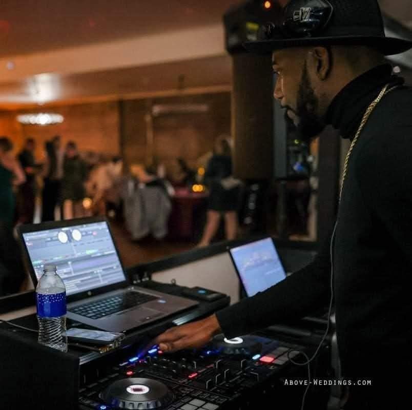 DJ in a black hat and turtleneck, mixing music at an event, laptop and mixer visible.