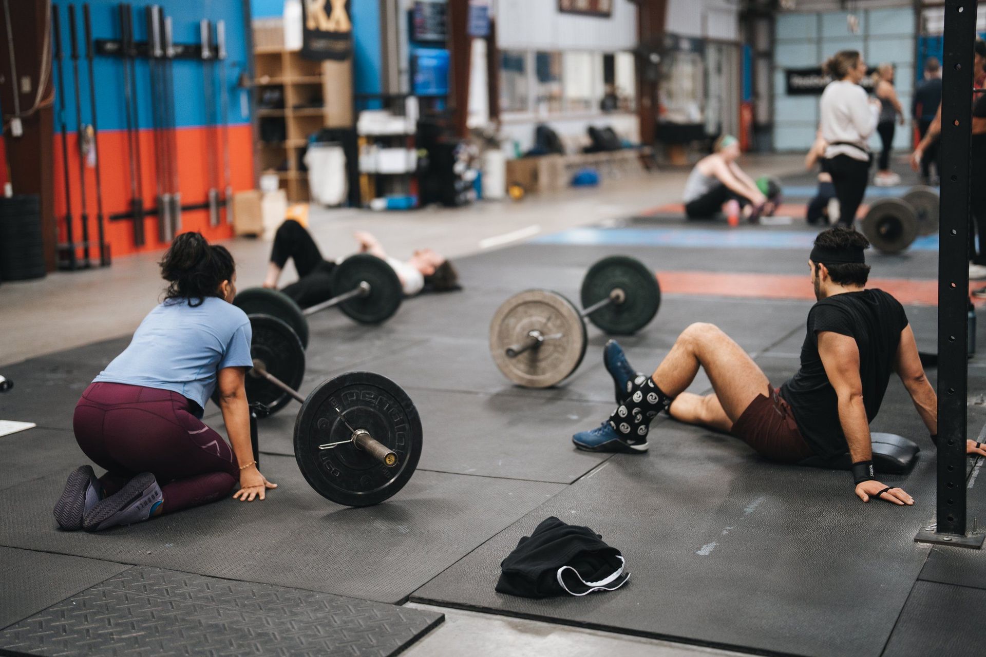 A group of people are sitting on the floor in a gym with barbells.