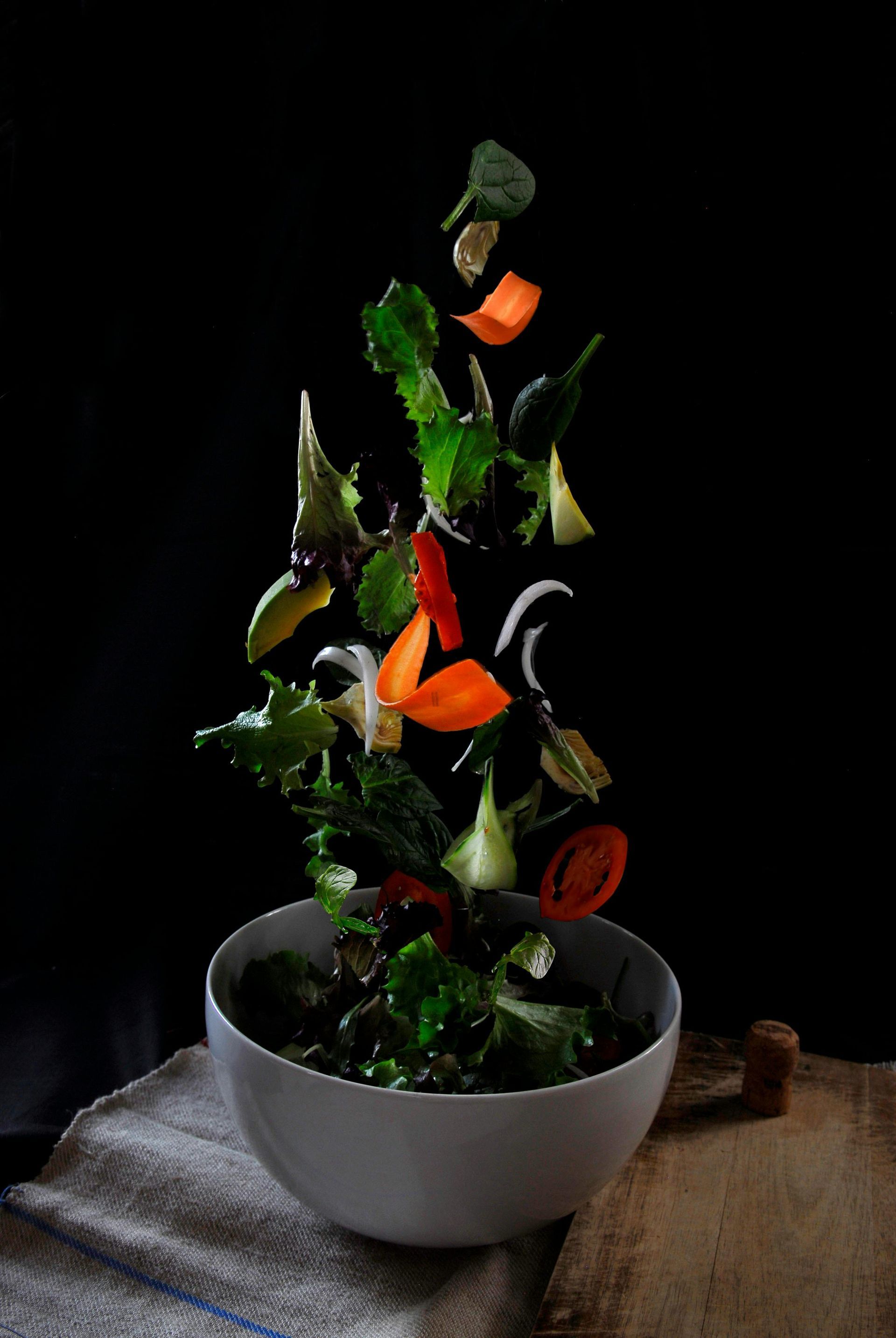 A bowl of salad with vegetables falling into it on a table.