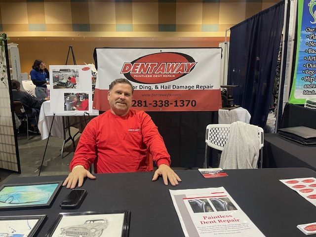 Man at DentAway booth, offering dent & hail repair services. Red shirt, black table, banner.
