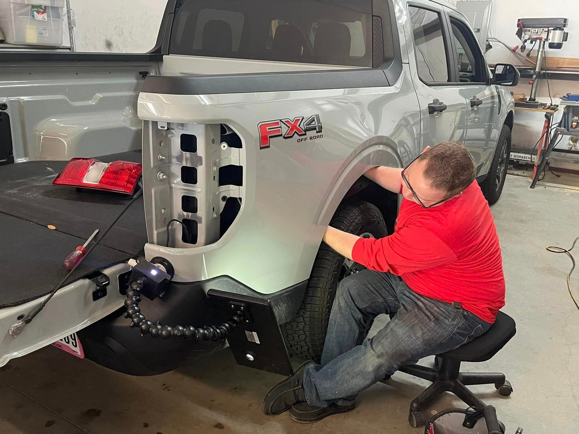 Man working on a gray pickup truck's fender. He's seated on a chair, wearing red shirt and jeans in a garage.
