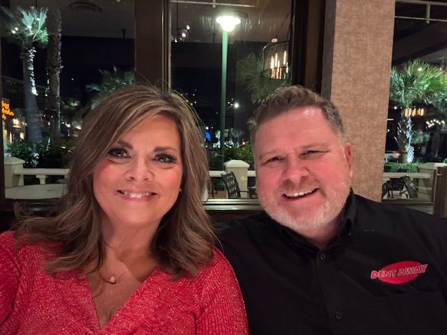 Woman and man smiling at a table, lit by streetlights. Woman wears red, man wears a black shirt.