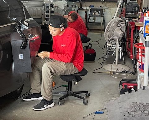Two men in red shirts repair a vehicle in a garage. One uses tools, the other works in the background.