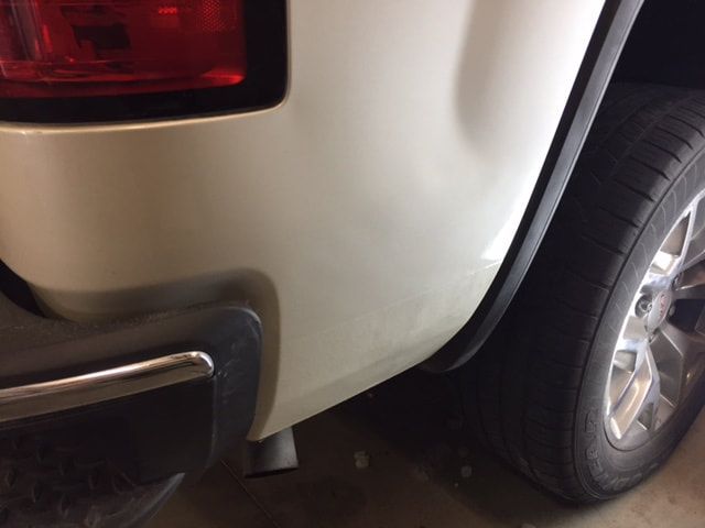 A close-up of a white pickup truck's rear bumper and tire; minor dent present.