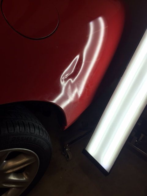Red car with dent in rear panel, lit by a dent repair light, near the tire.