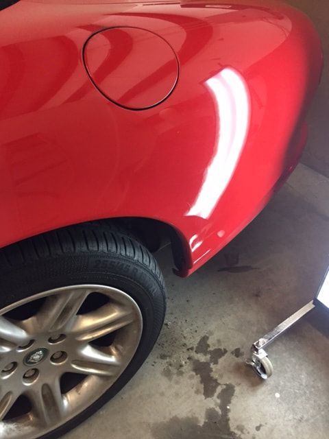 Red car's rear side with tire and fuel door visible. Garage setting with shiny paint and light reflections.
