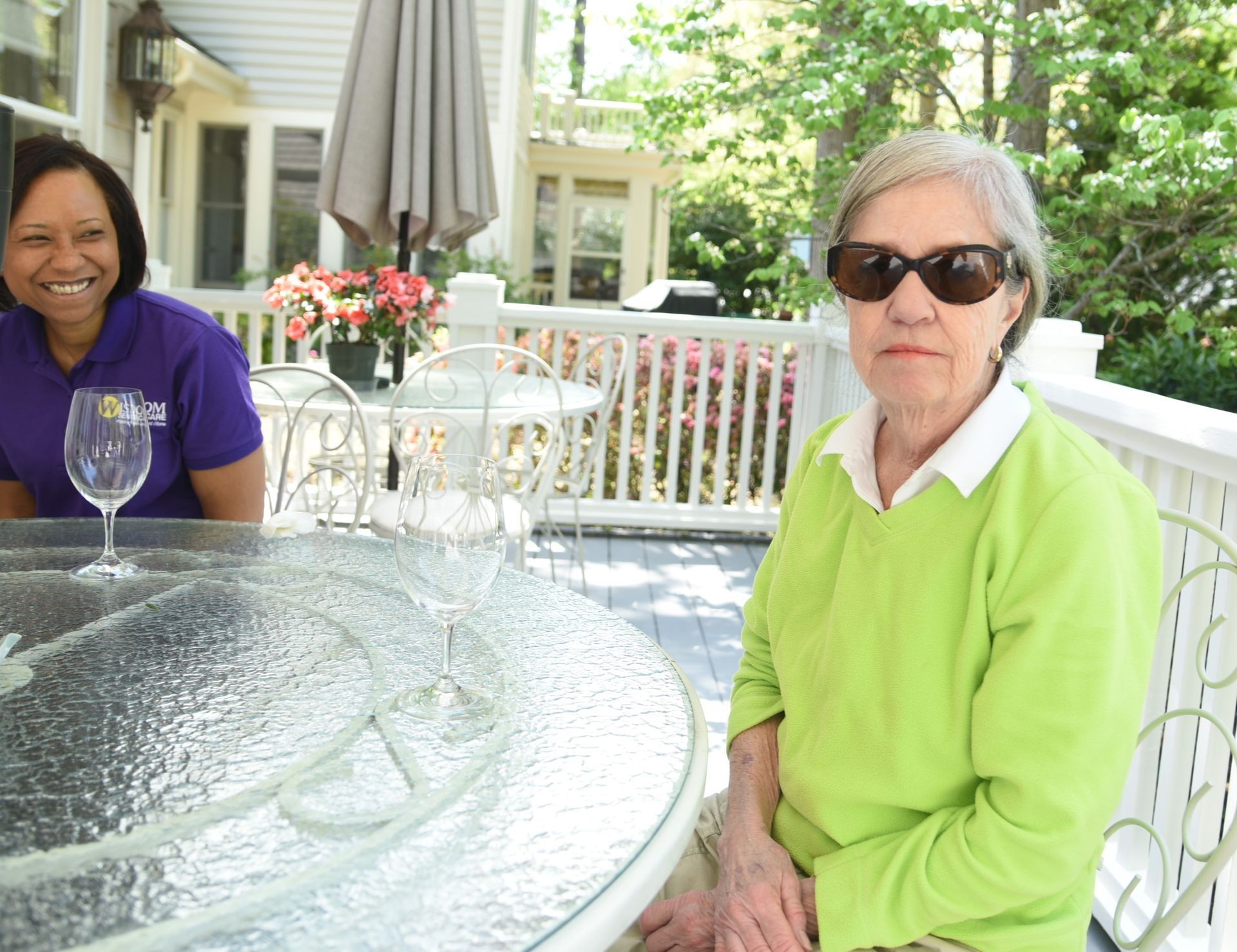 Woman in sunglasses and lime green sweater sitting at a glass table on a sunny porch; another woman smiles in background.