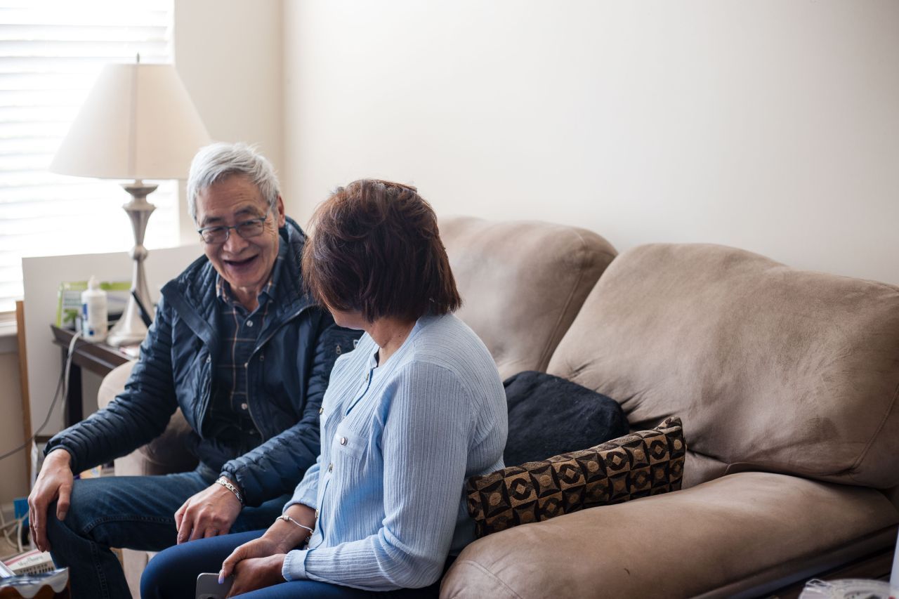 Two people laughing on a couch in a well-lit living room; man in a vest, woman in a light blue shirt.