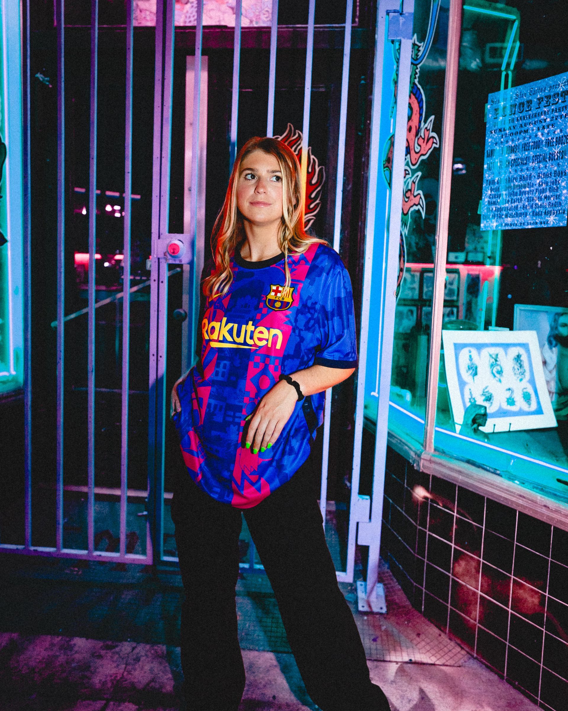 Woman wearing Barcelona jersey, standing in front of a gate at night with neon lights.