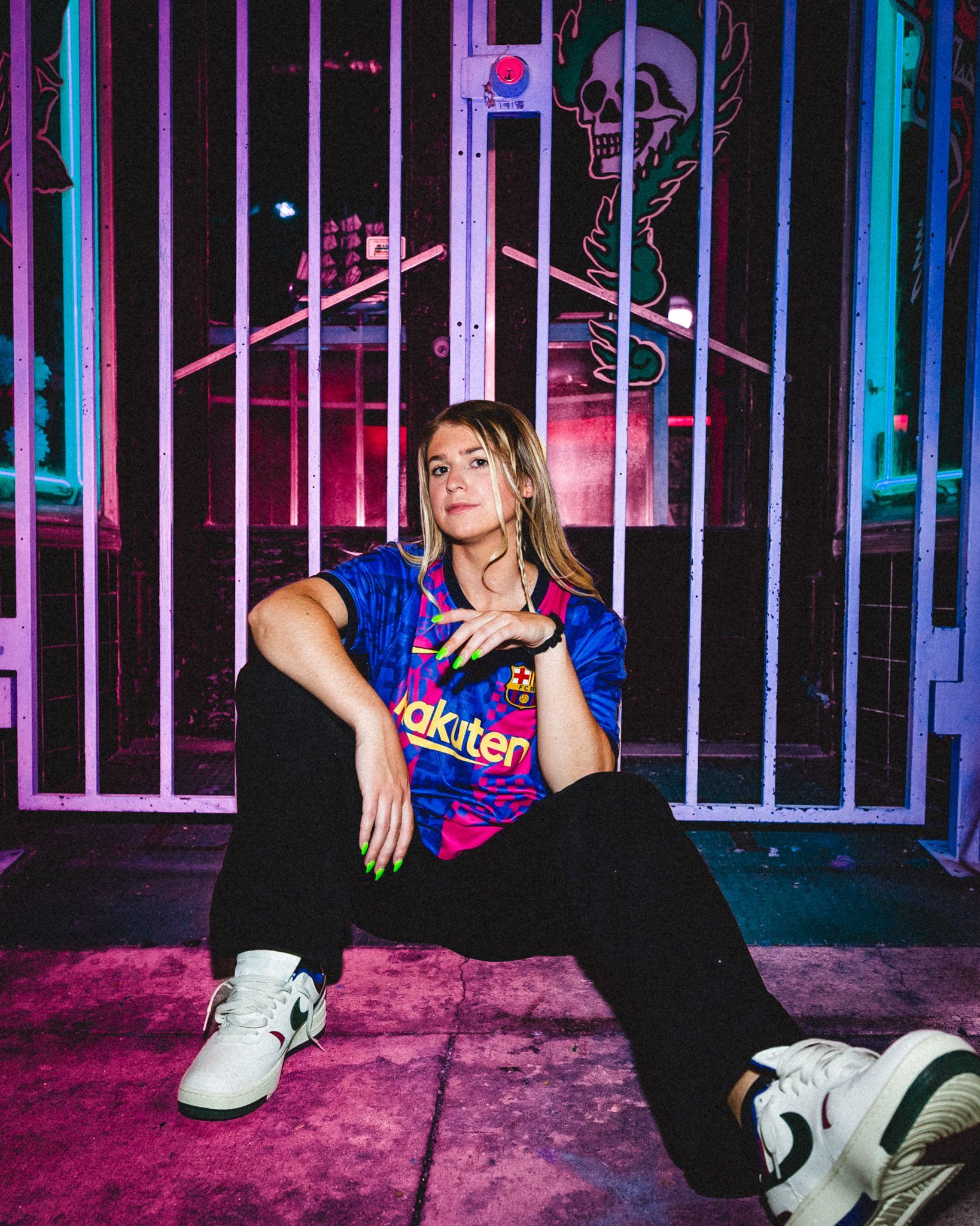 Woman in FC Barcelona jersey and sneakers, sitting in front of a gate, at night. Neon lights.