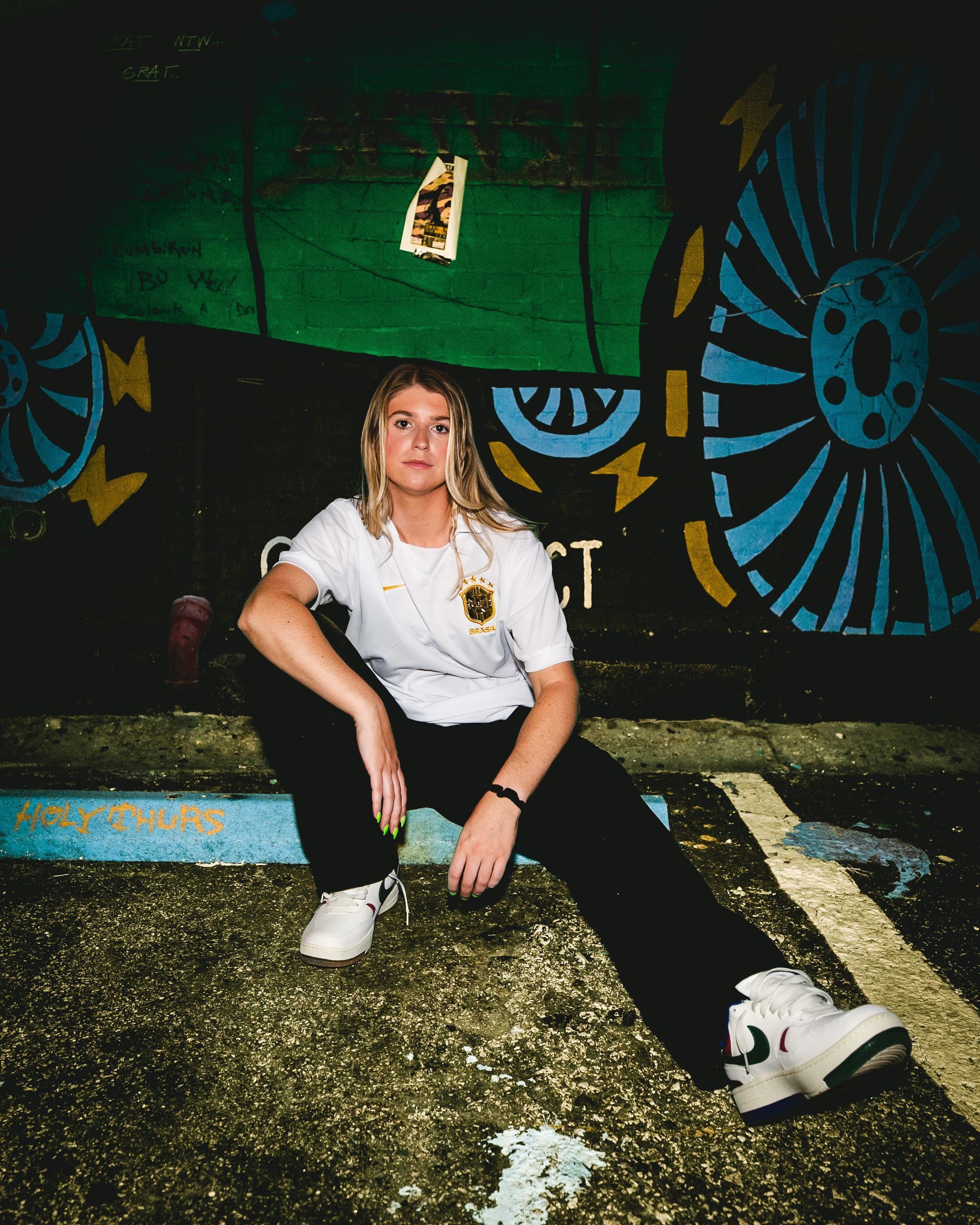 Woman in white t-shirt and black pants, crouching in front of a mural with blue and yellow accents.