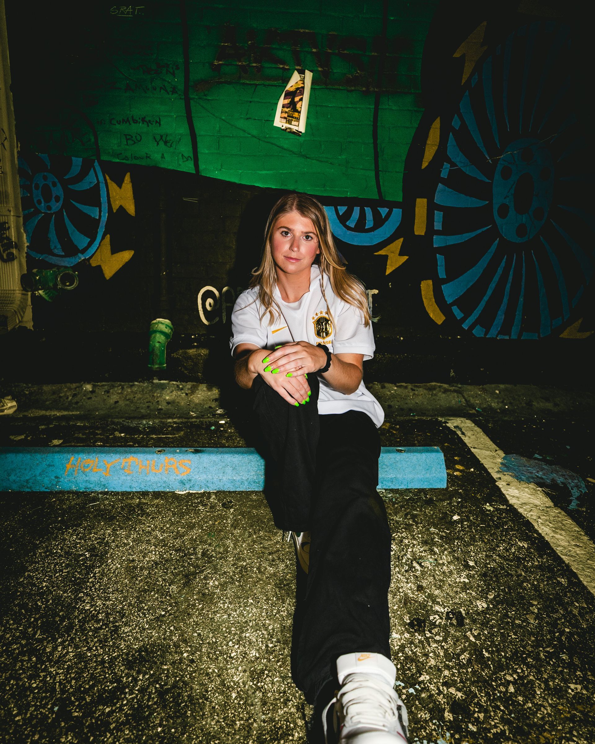 A person sits on a blue curb in front of a mural. The person is wearing white and black clothing.