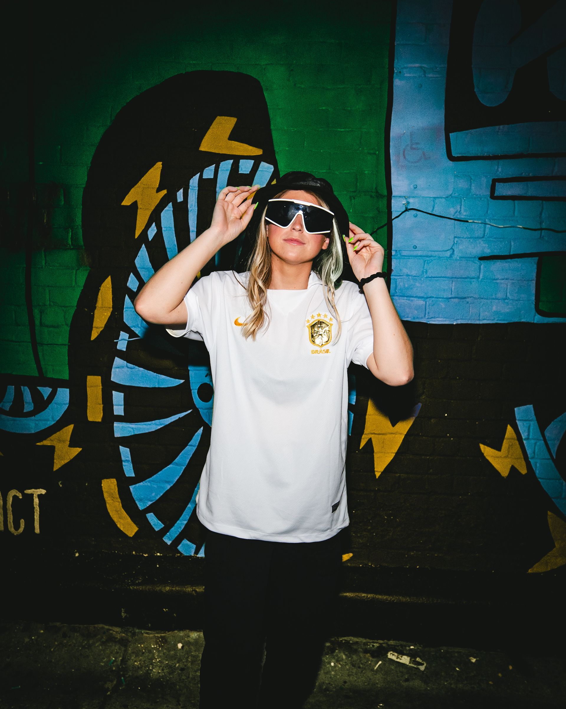 Person in sunglasses and a white shirt with a logo, standing in front of a colorful graffiti wall.