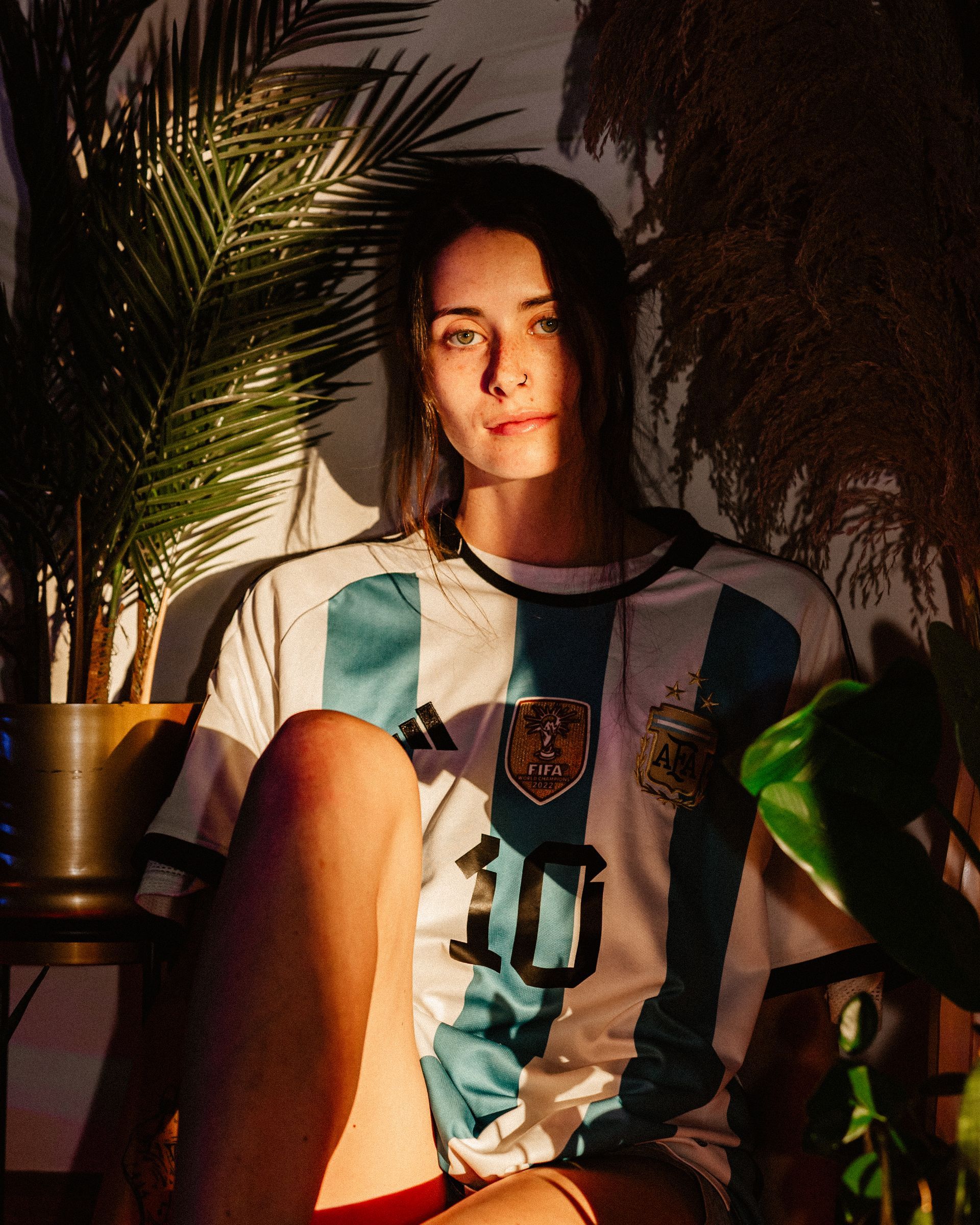 Woman wearing Argentina soccer jersey, sitting near plants, lit by sunlight.