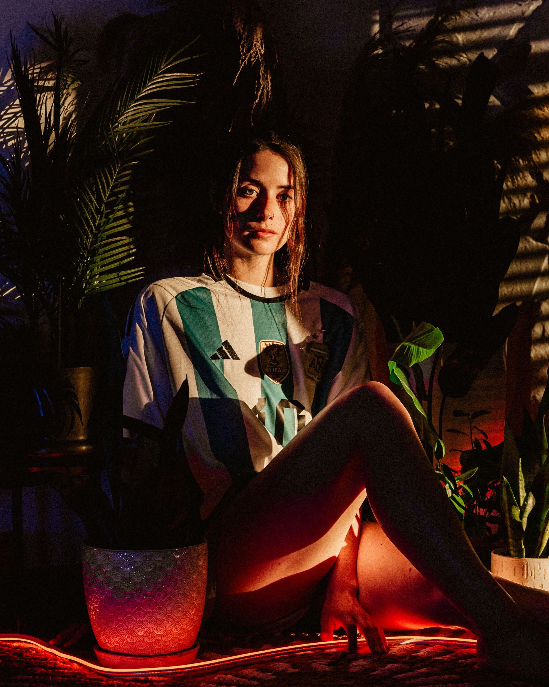 Woman wearing a blue and white Argentina soccer jersey, sitting amongst plants, lit by red light.