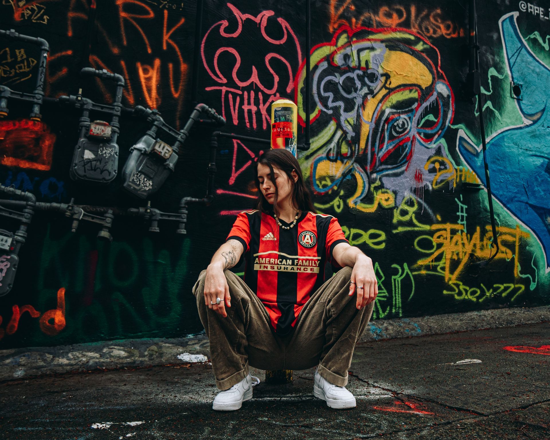 Person squatting in front of graffiti wall, wearing soccer jersey and patterned pants.