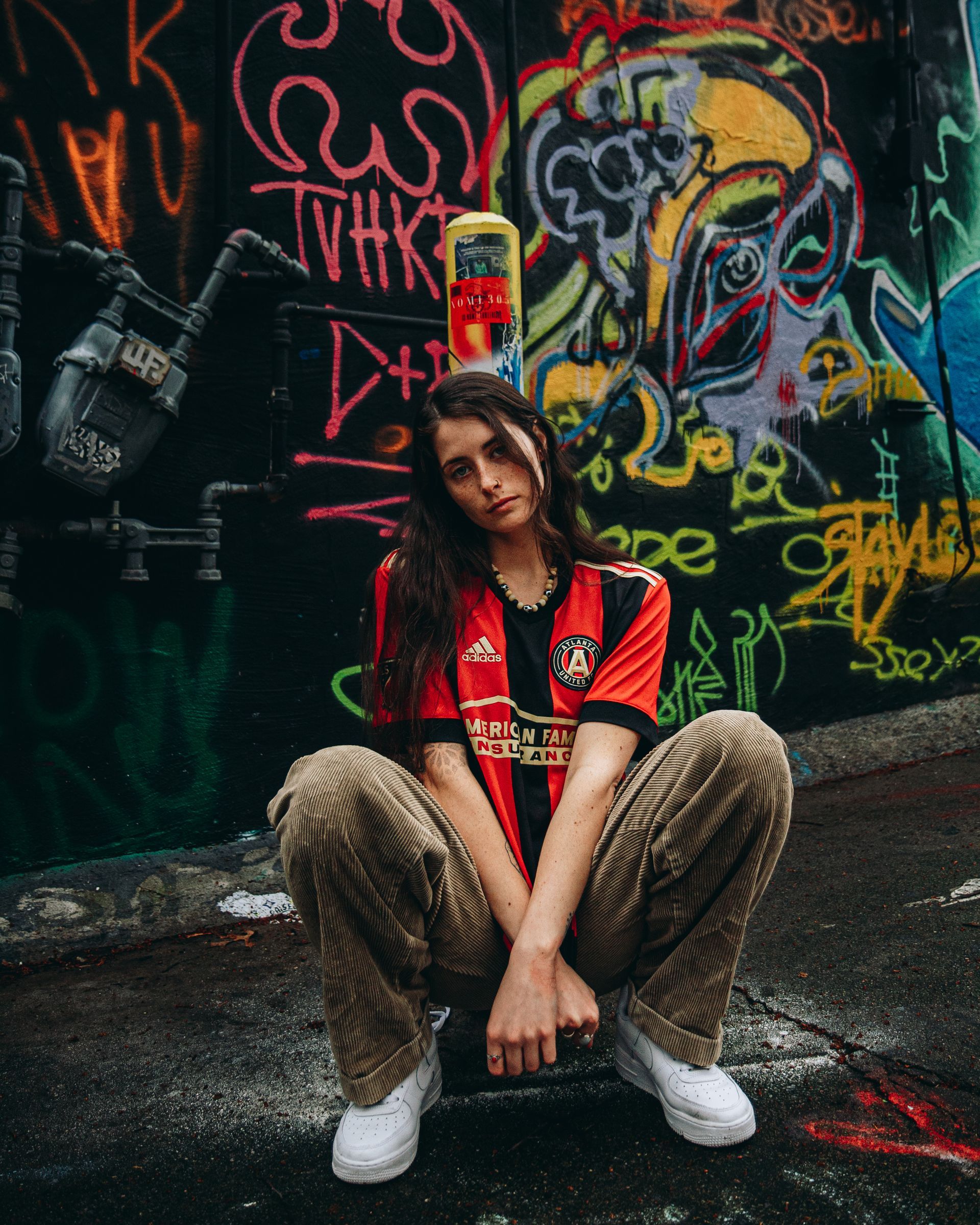 Person squatting, wearing a red jersey and patterned pants, in front of a colorful graffiti wall.