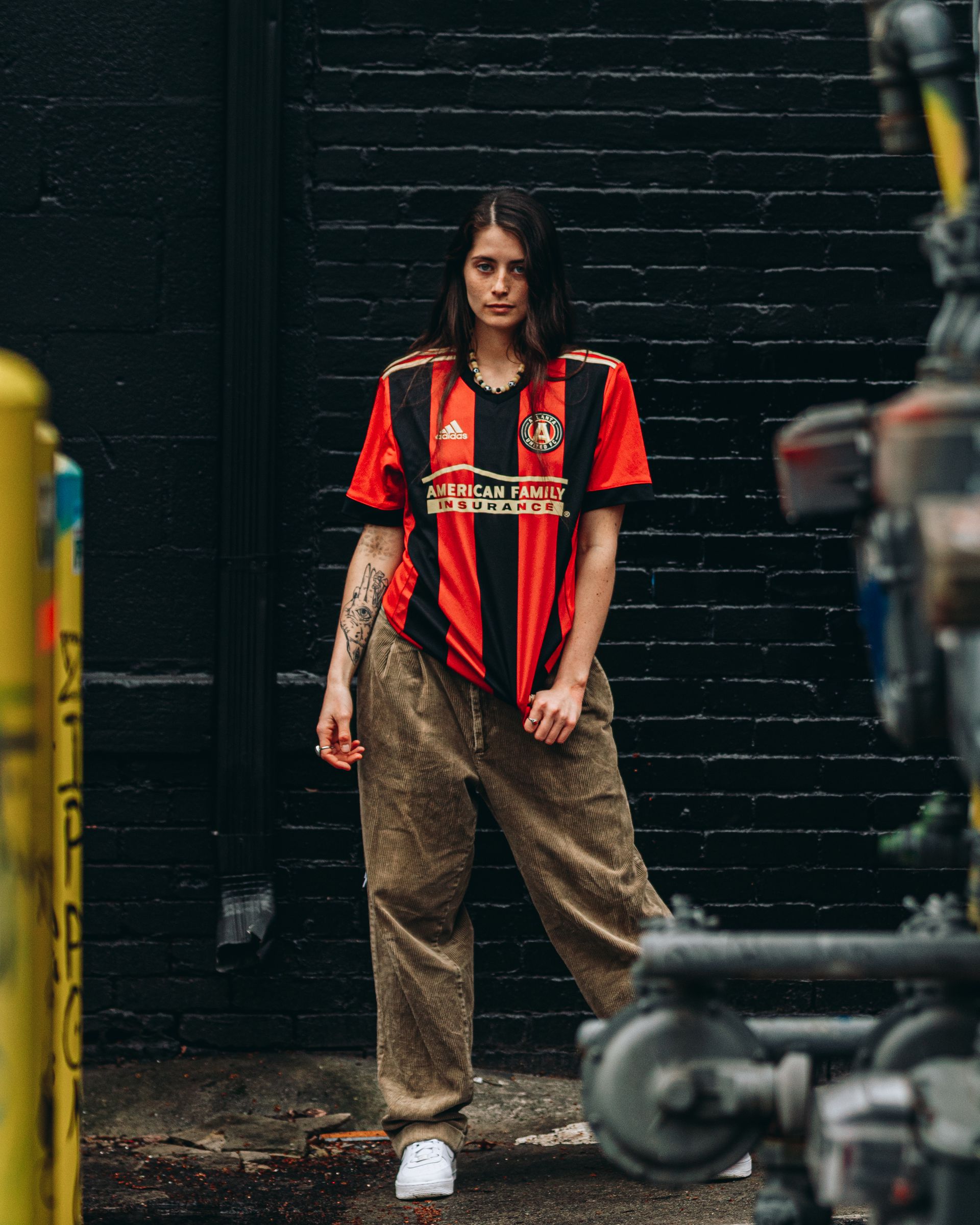 Person wearing a black and red soccer jersey and brown pants stands in front of a dark brick wall.