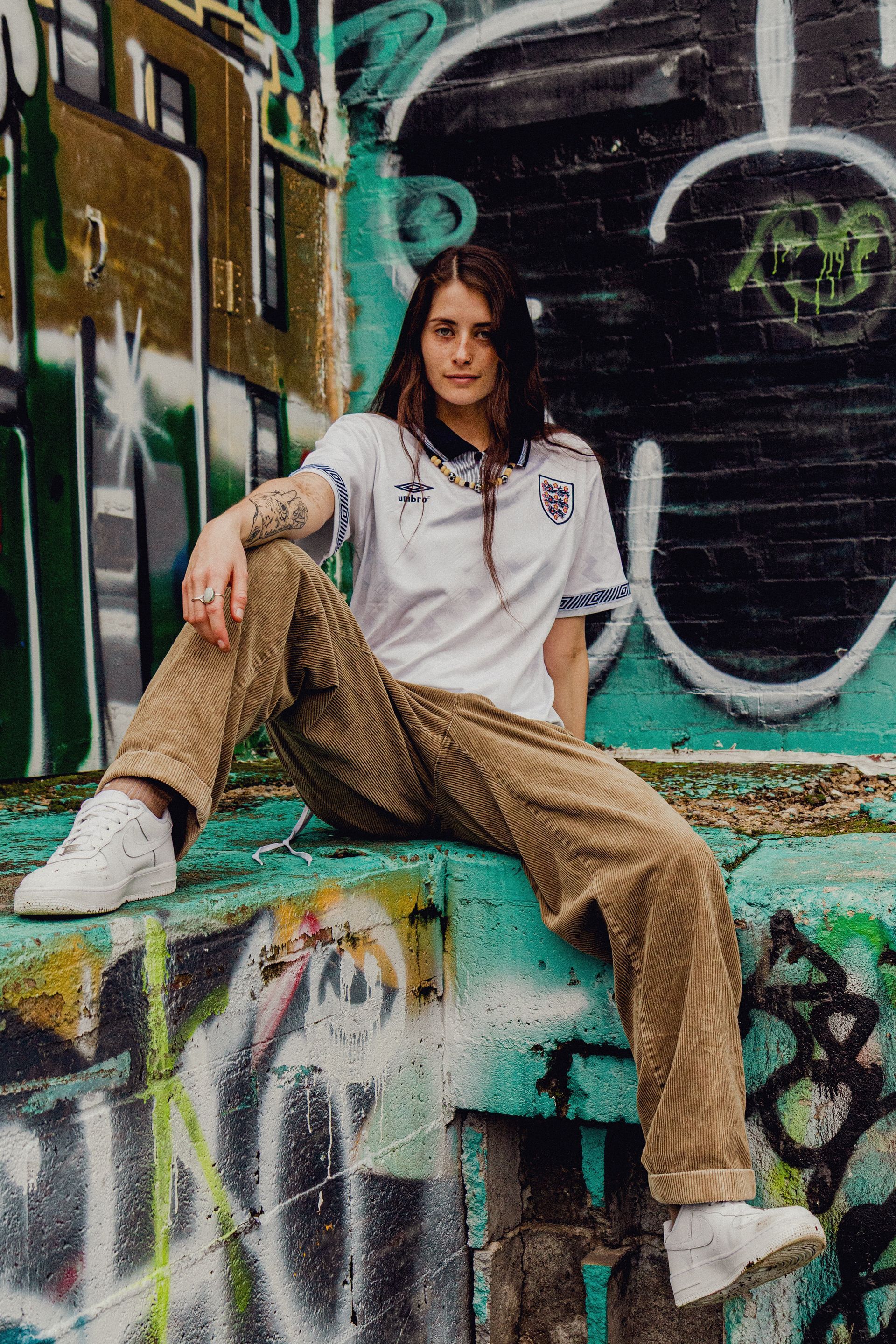 Person wearing a soccer jersey and corduroy pants sits on a graffiti-covered ledge.