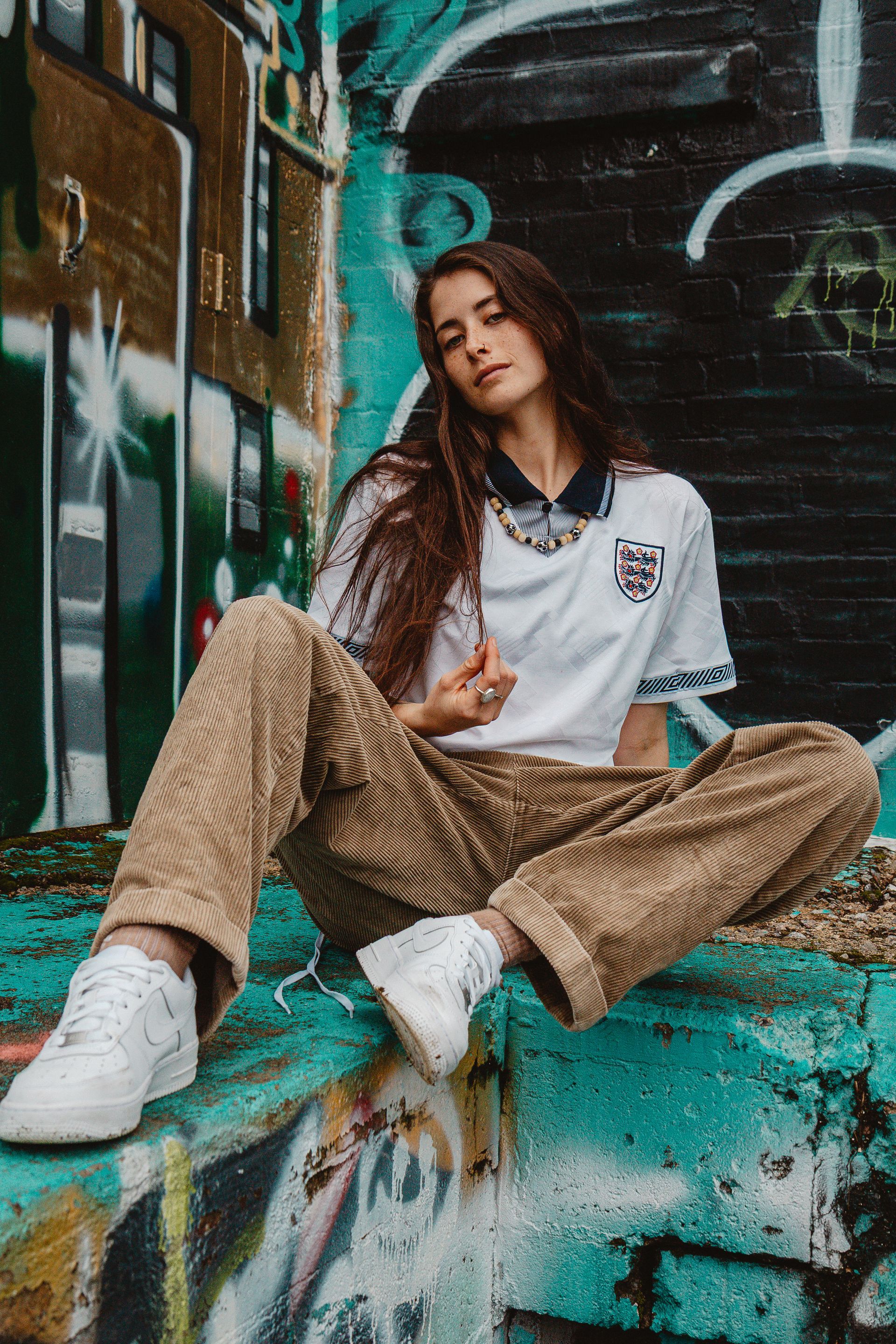 Woman in white England jersey and corduroy pants sits by graffiti-covered wall, gazing at the viewer.