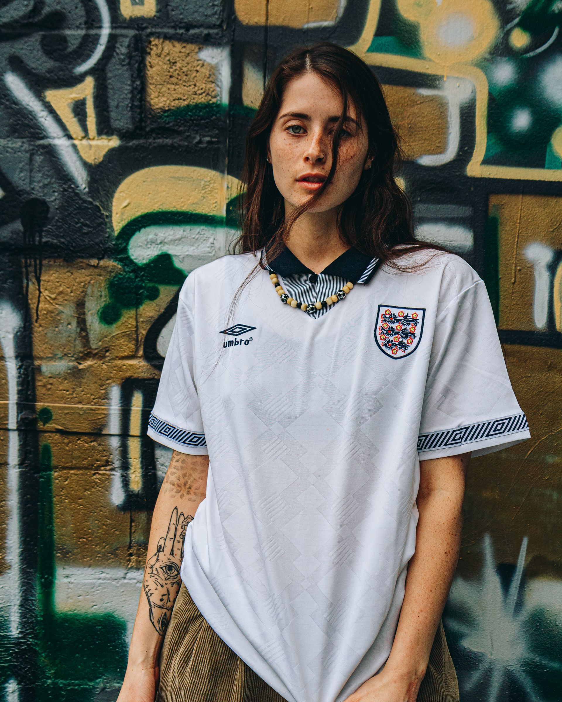 Woman in white England soccer jersey stands against a graffiti wall.
