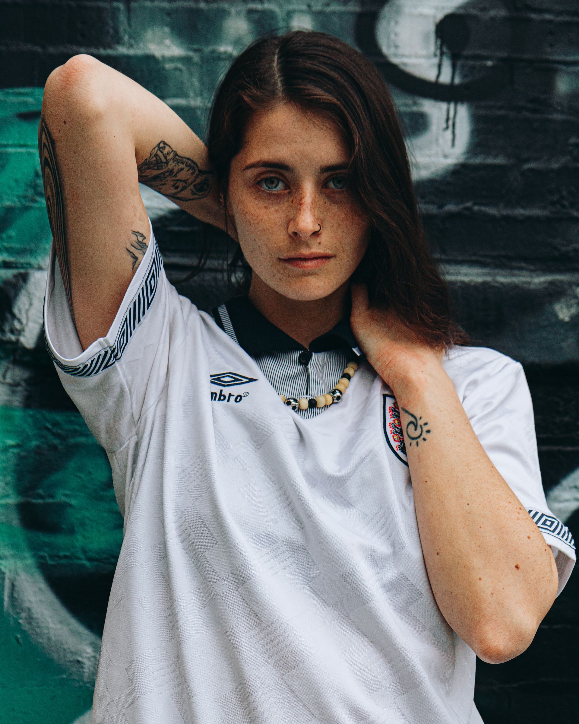 Woman in white soccer jersey with tattoos, hands behind head, against a graffiti wall.