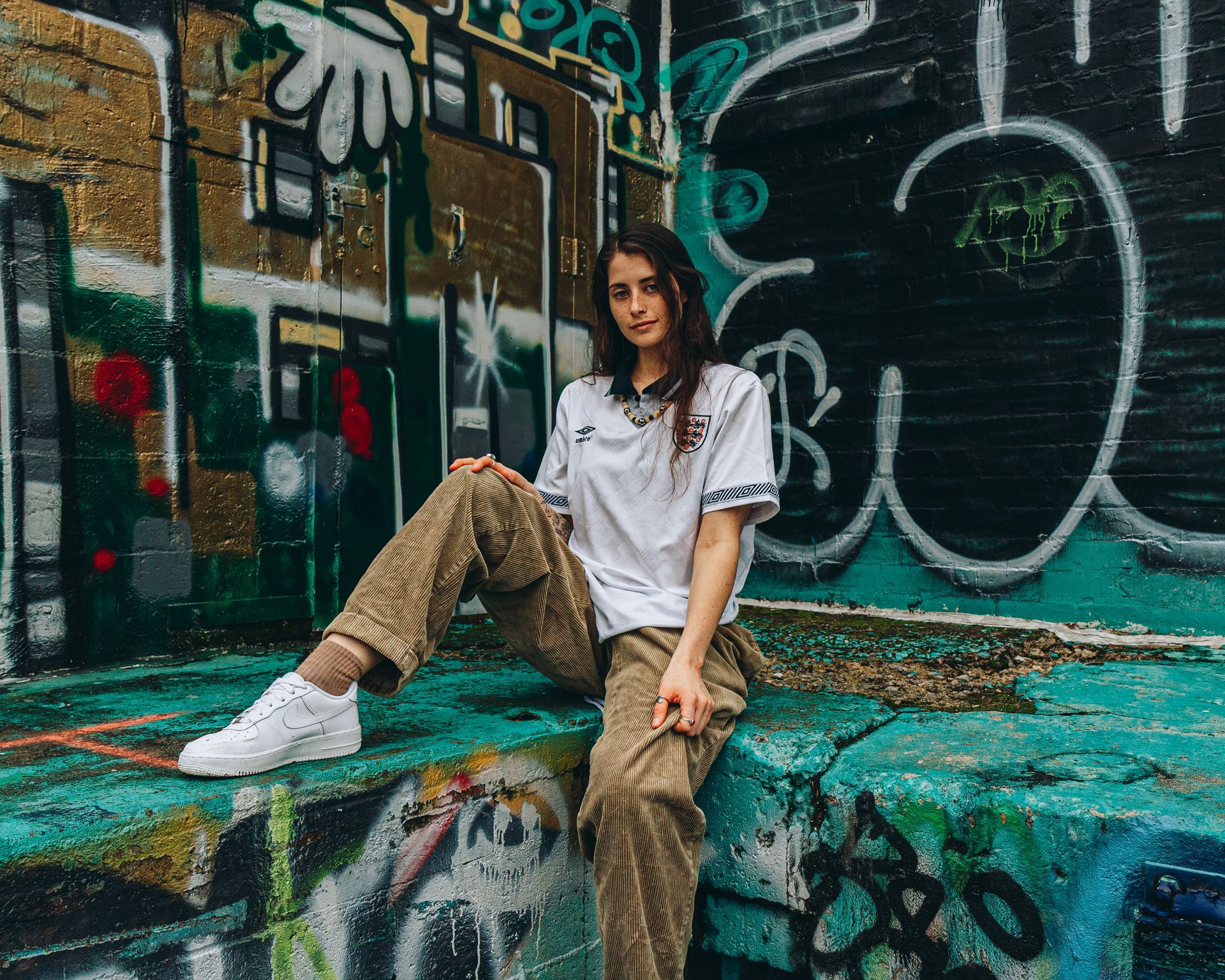Woman in white top and corduroy pants sits by graffiti-covered wall, posing.