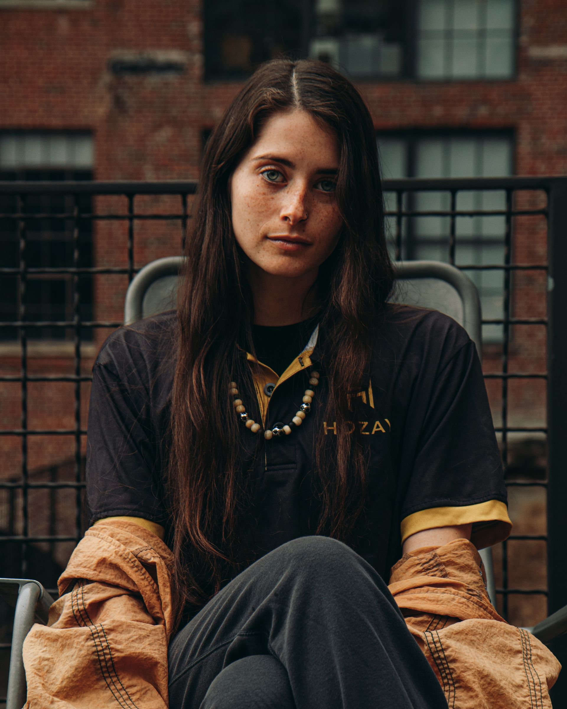 Woman with long dark hair, wearing a black shirt and seated outside with brick building background.