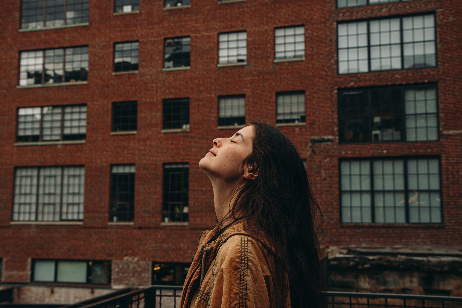 Woman with eyes closed, facing upwards, against a brick building backdrop. Sunlight on her face.