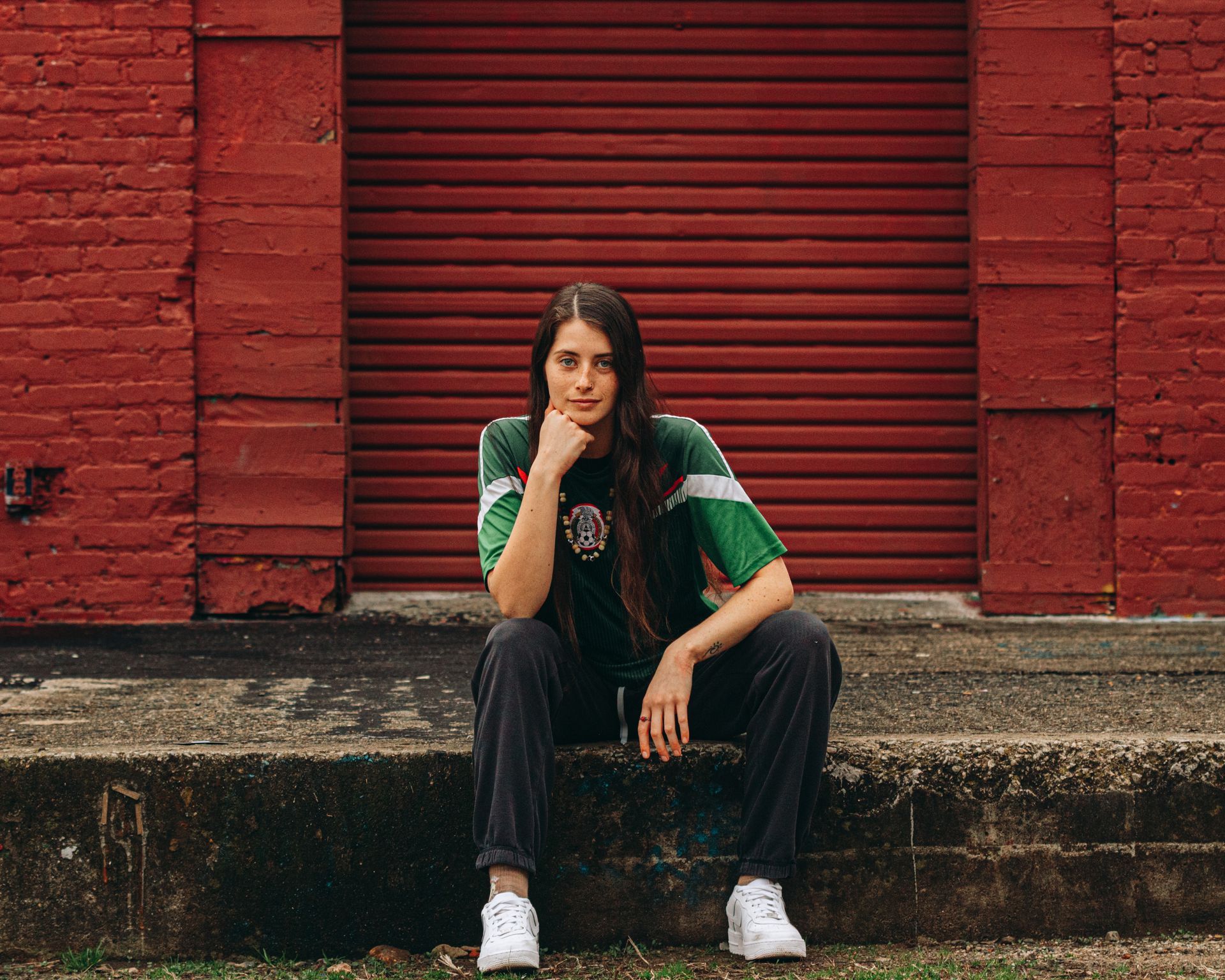 Person sitting on steps in front of a red, shuttered building, wearing a green shirt, dark pants, and white sneakers.