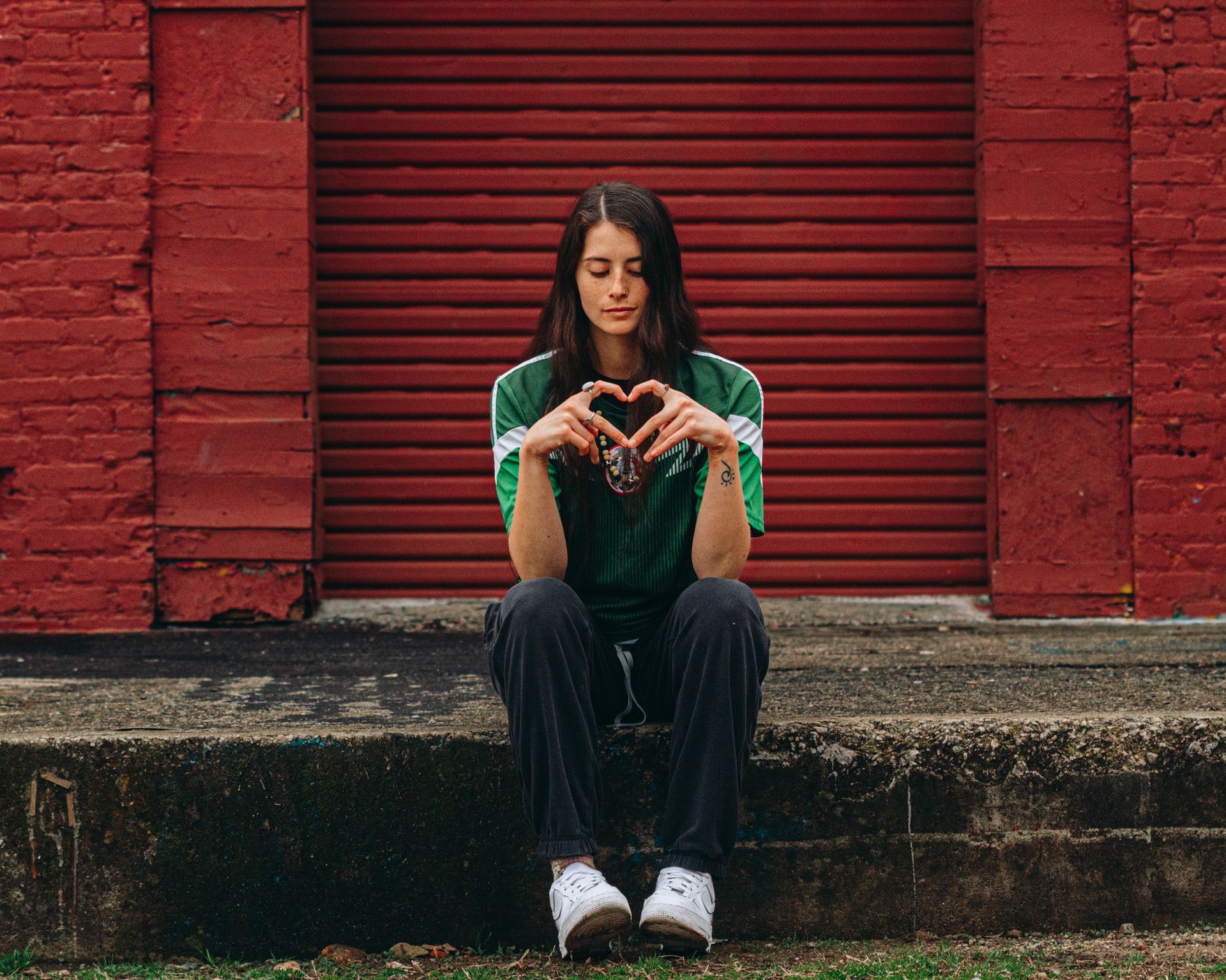 Woman in green shirt, forming a heart with hands, sits on concrete step against red brick wall.