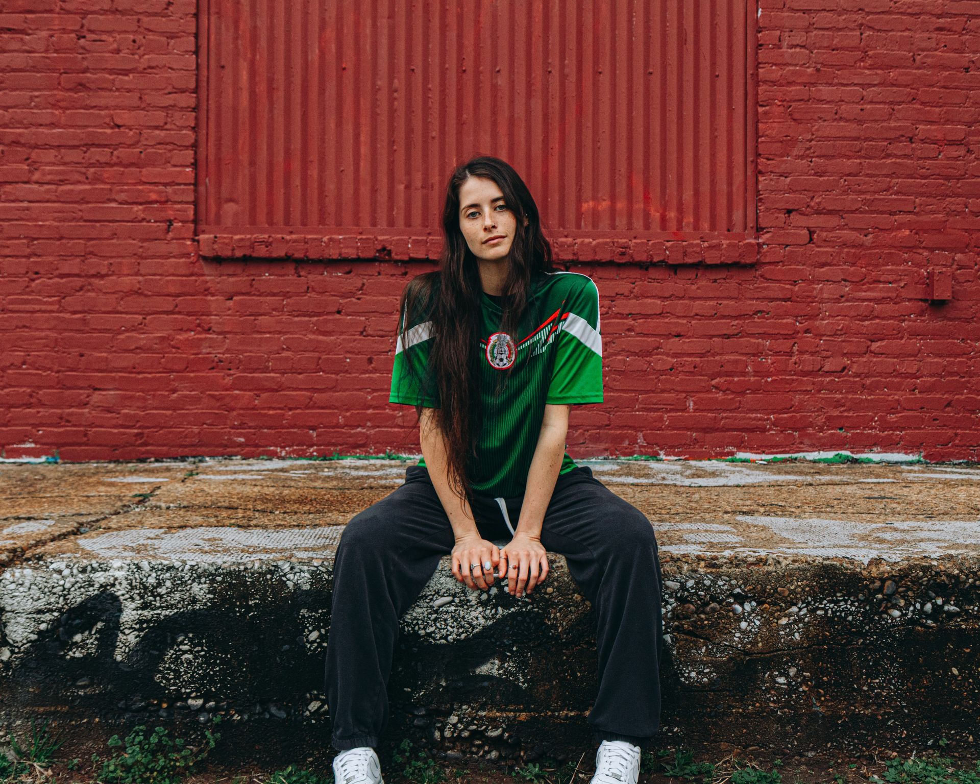 Woman seated outdoors wearing a green soccer jersey and gray pants. Leaning against a red wall.