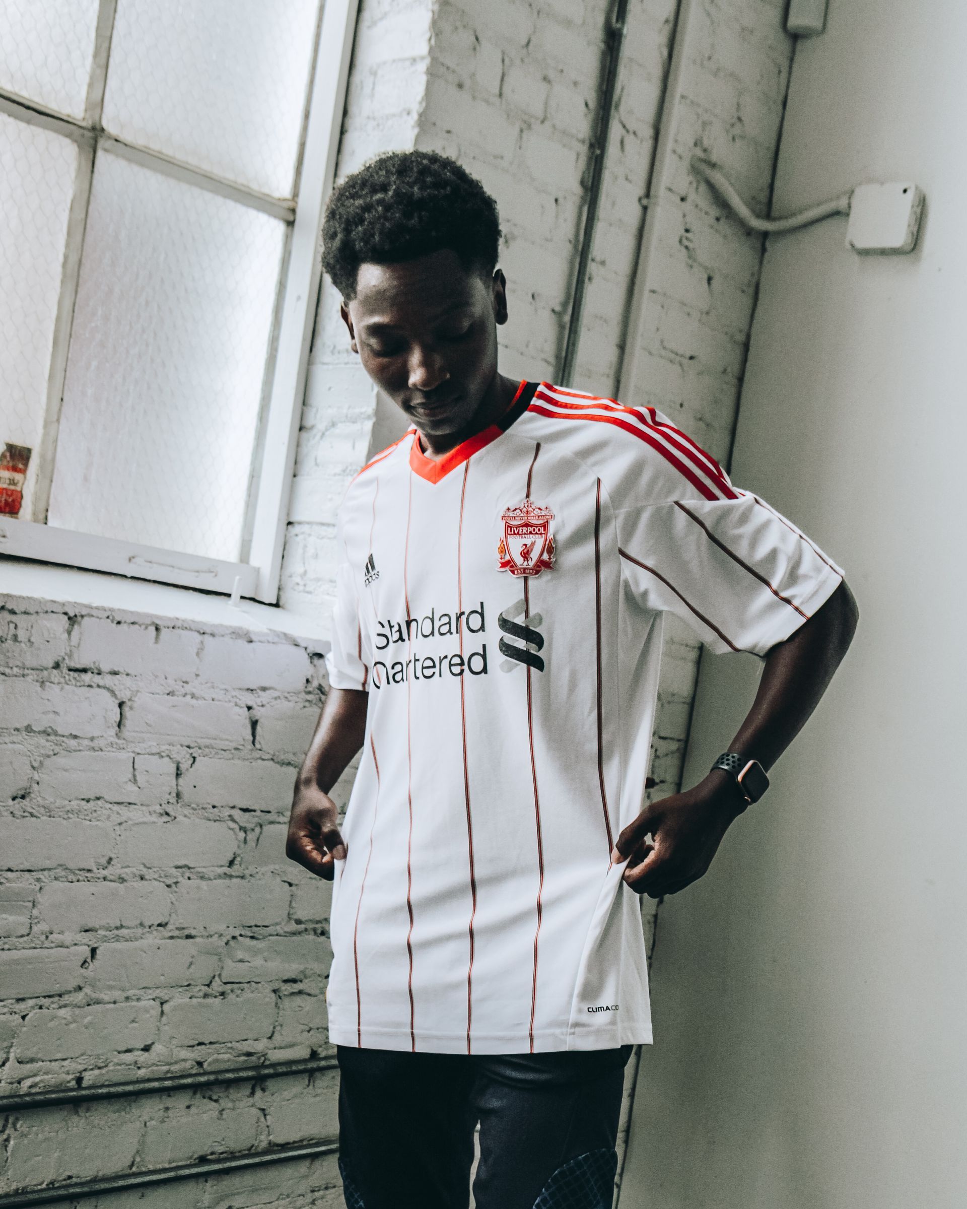 Man in white Liverpool jersey, red stripes, standing by a window.