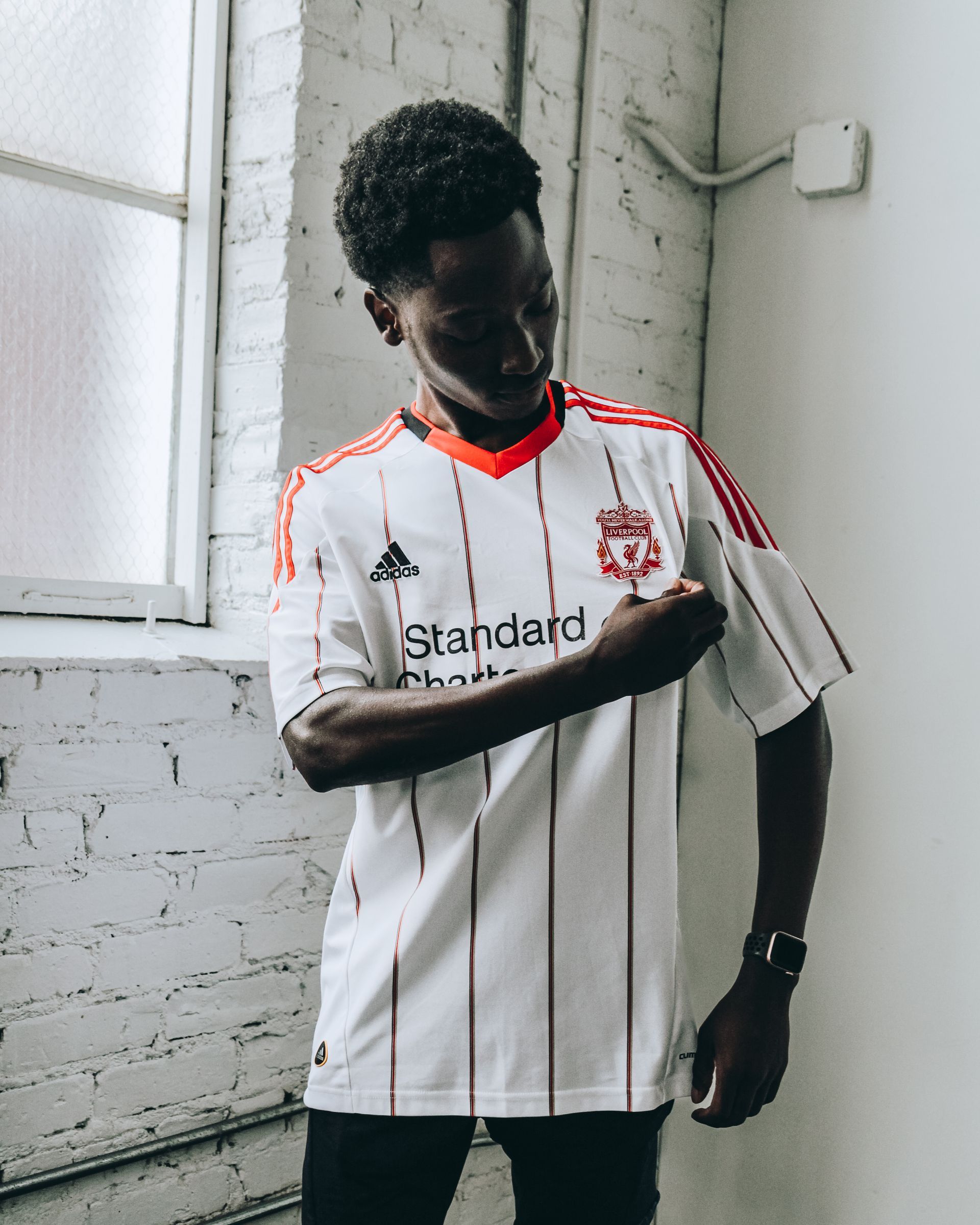 Person wearing a white Liverpool soccer jersey with red accents, in a brick-walled room, touching the jersey.