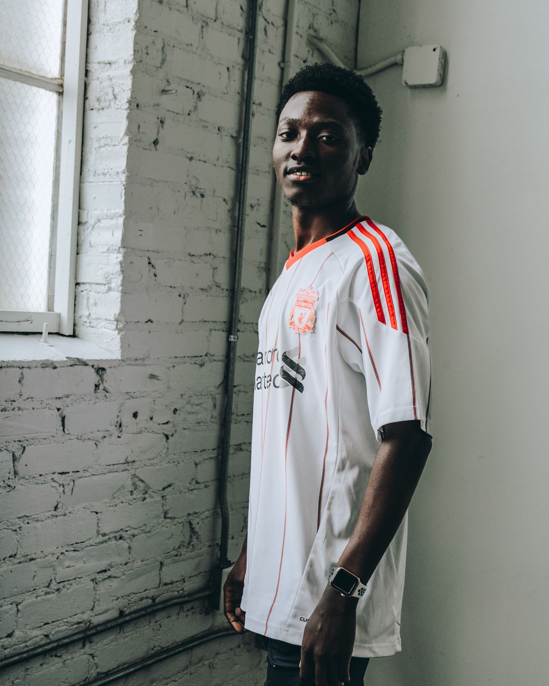 Man in white soccer jersey smiles near a brick wall and window.