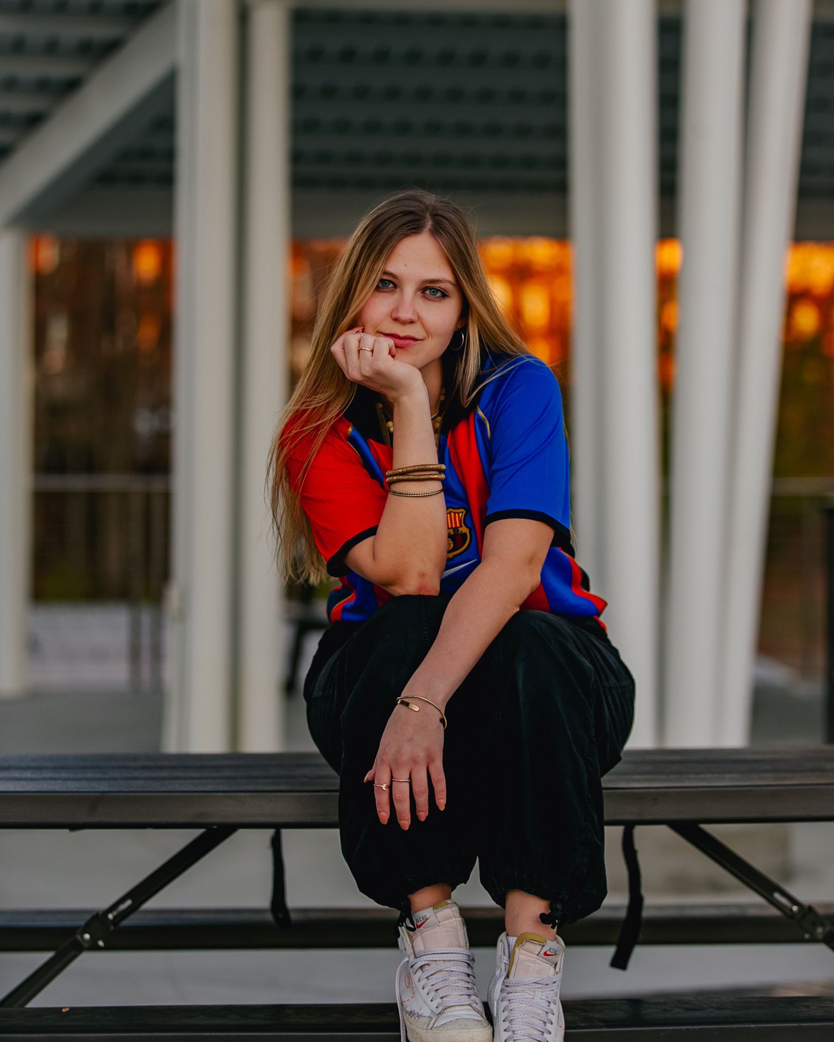 Woman in a Barcelona jersey sits on a bench, resting her chin on her hand, with a blurred background of architectural support beams.