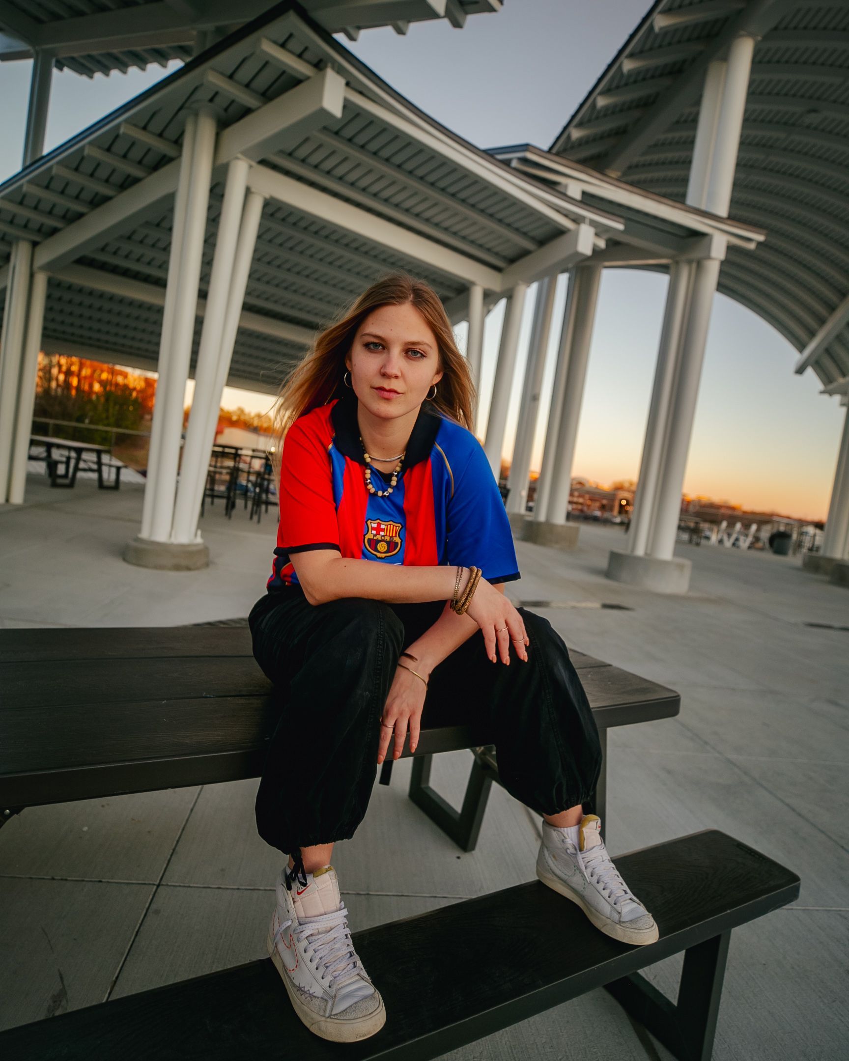 Person in a blue, red, and yellow shirt, black pants, and white sneakers, sitting on a bench in a pavilion.