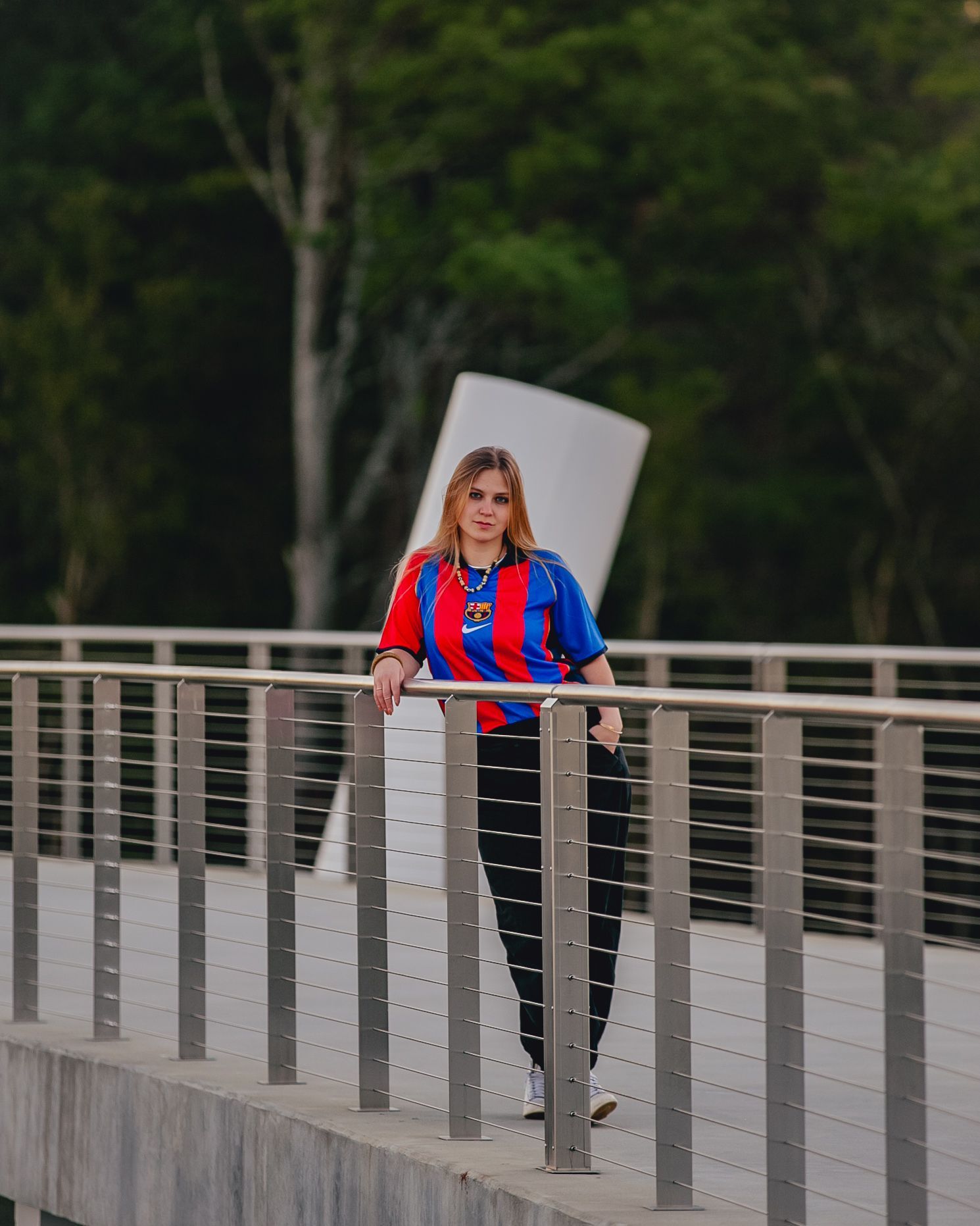 Woman in red, blue, and white jersey, black pants, on a bridge, holding onto a railing, with a modern sculpture in the background.