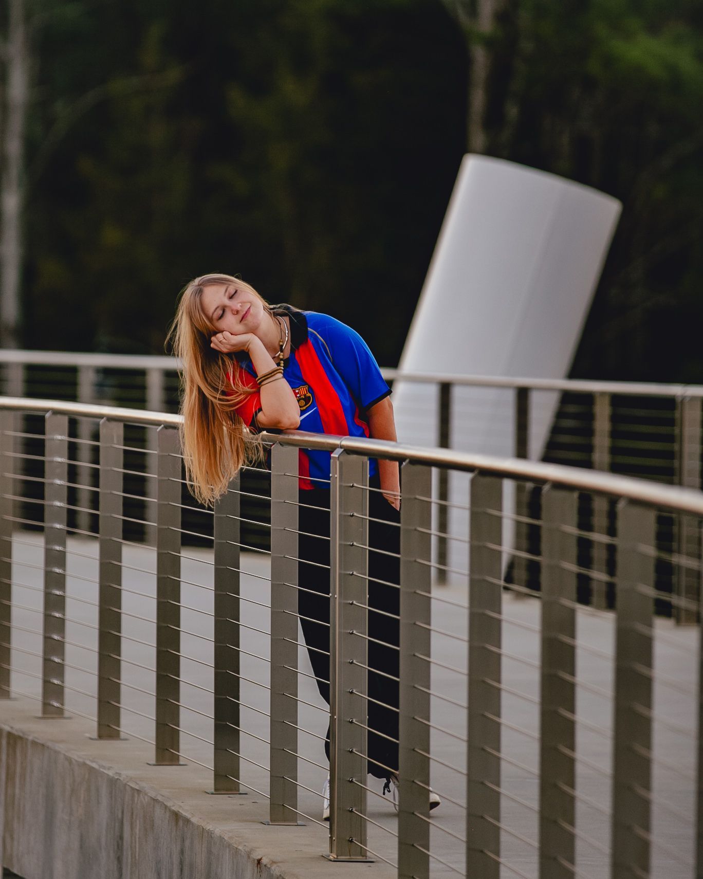 Woman leaning on railing, smiling, wearing a blue and red soccer jersey, black pants, on a bridge, white modern art in background.