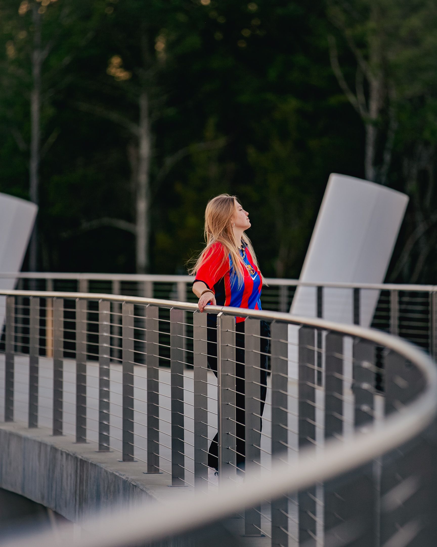 Woman wearing a red and blue shirt, looking up from a curved bridge with a metal railing. Trees and white structures in the background.