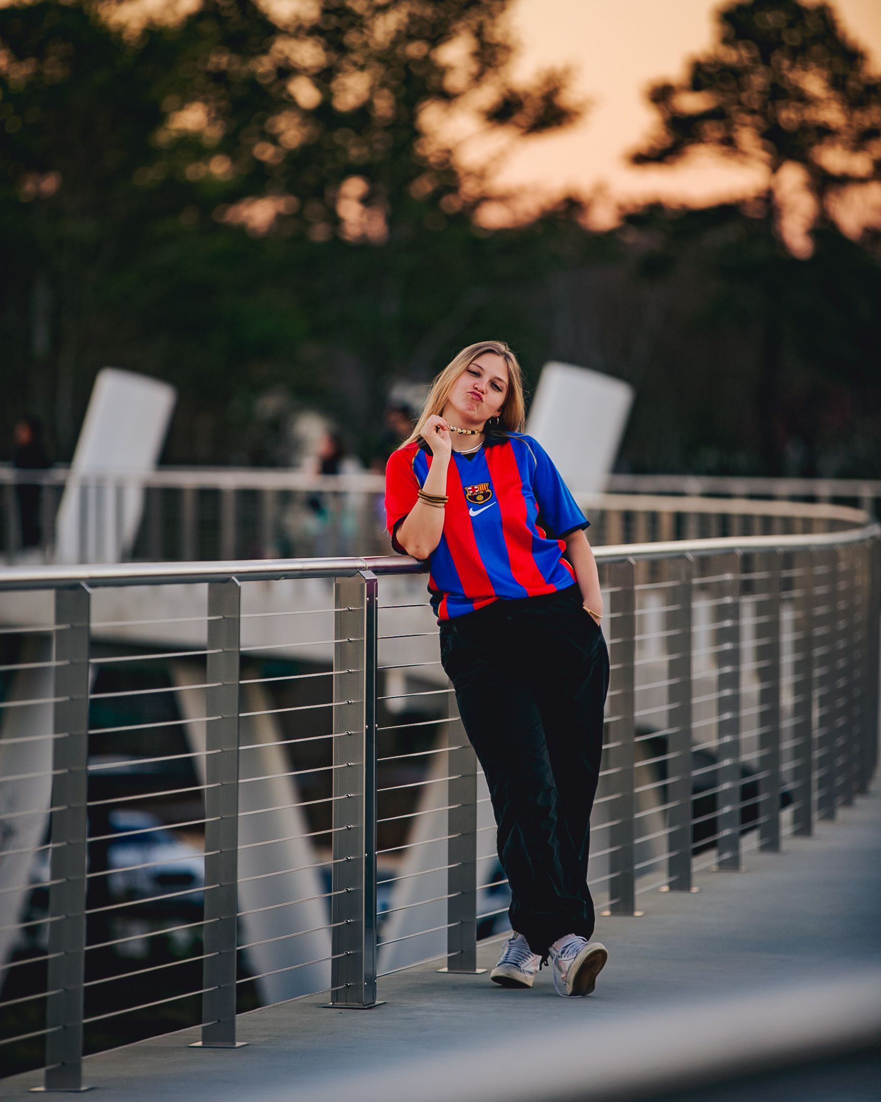 Woman leans on railing, wearing a blue and red striped jersey and black pants, outdoors at dusk.
