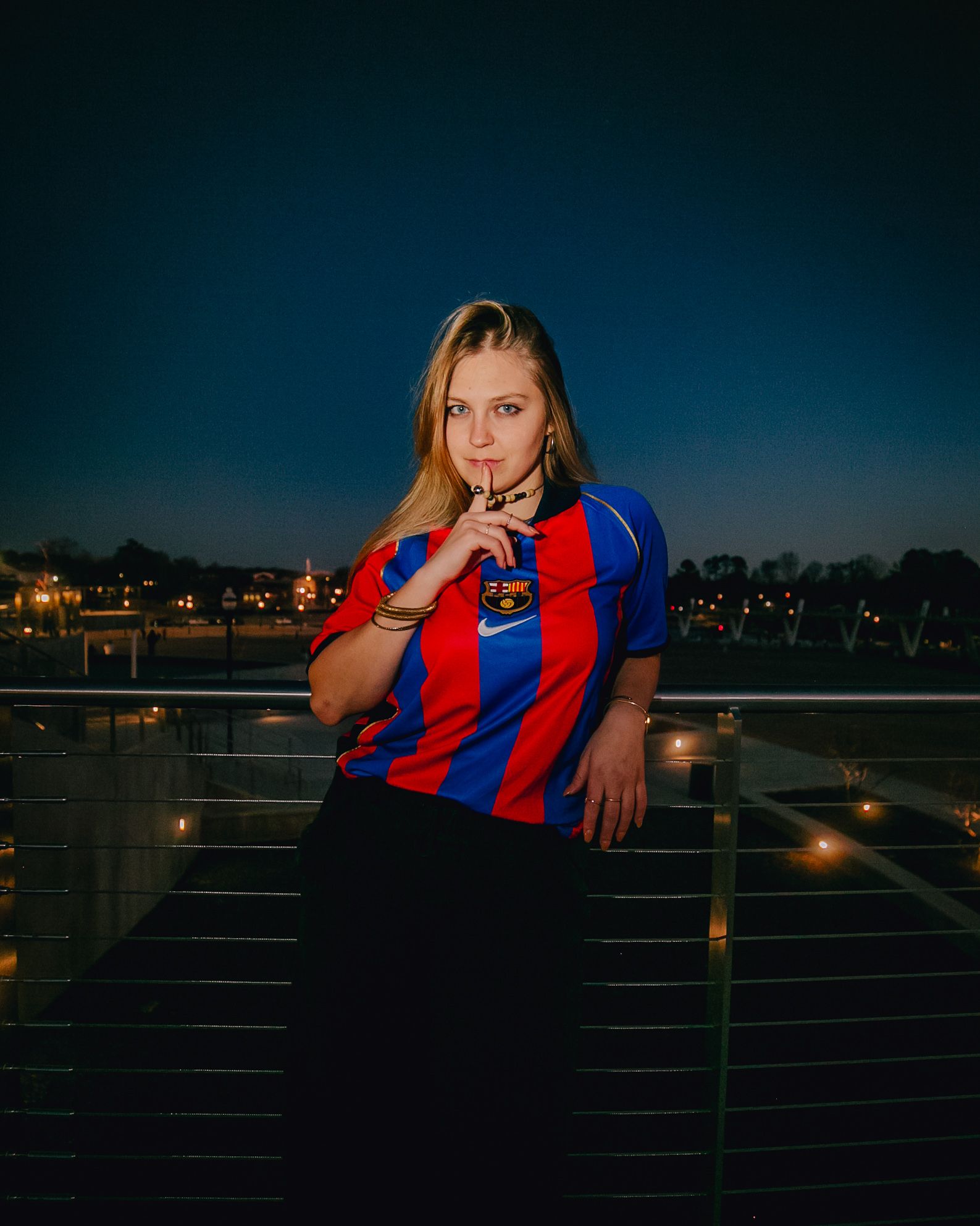 Woman wearing Barcelona jersey, standing outdoors at night, holding a cross, dark background.