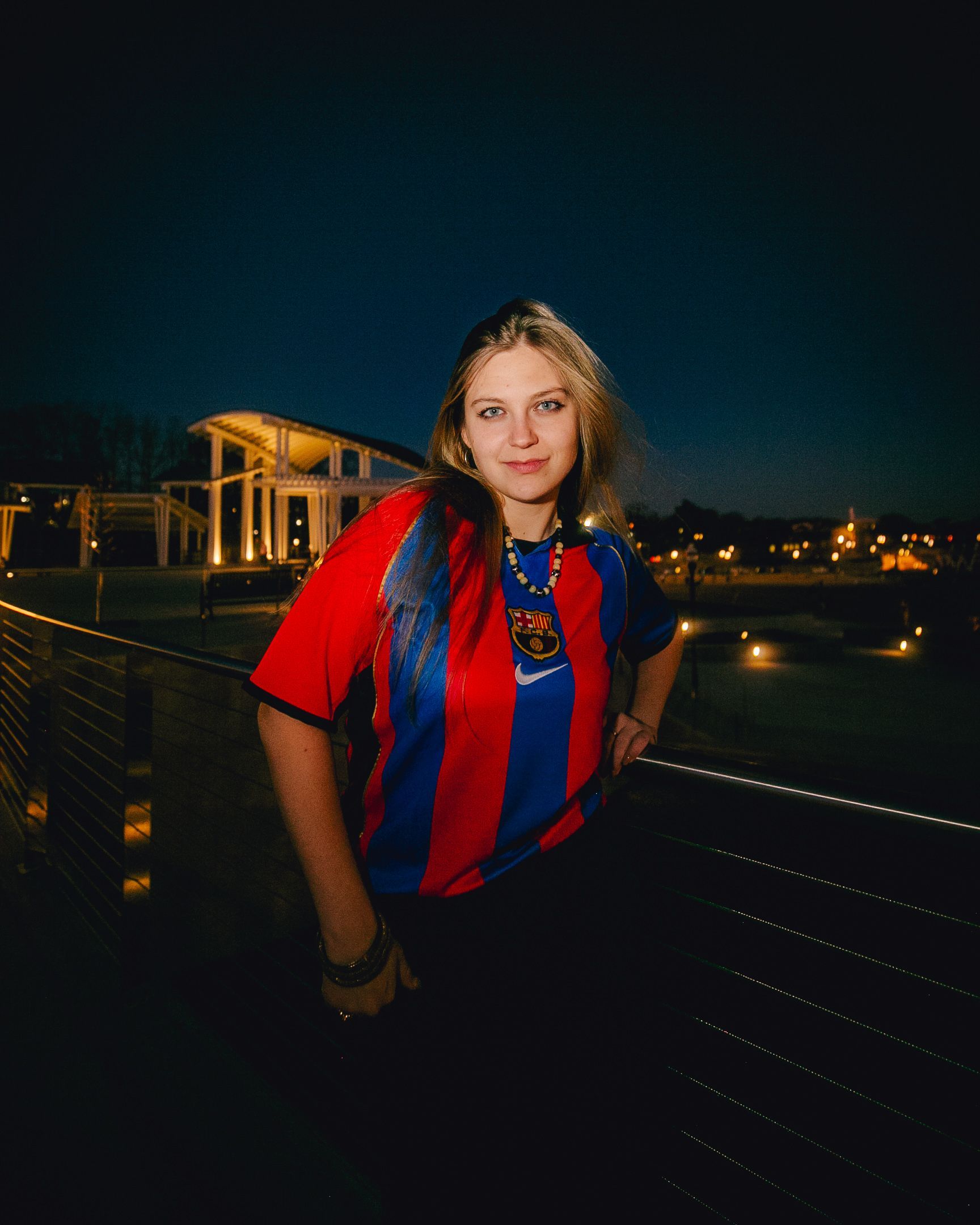 Woman in striped red/blue shirt stands on a deck at night with a building in the background.